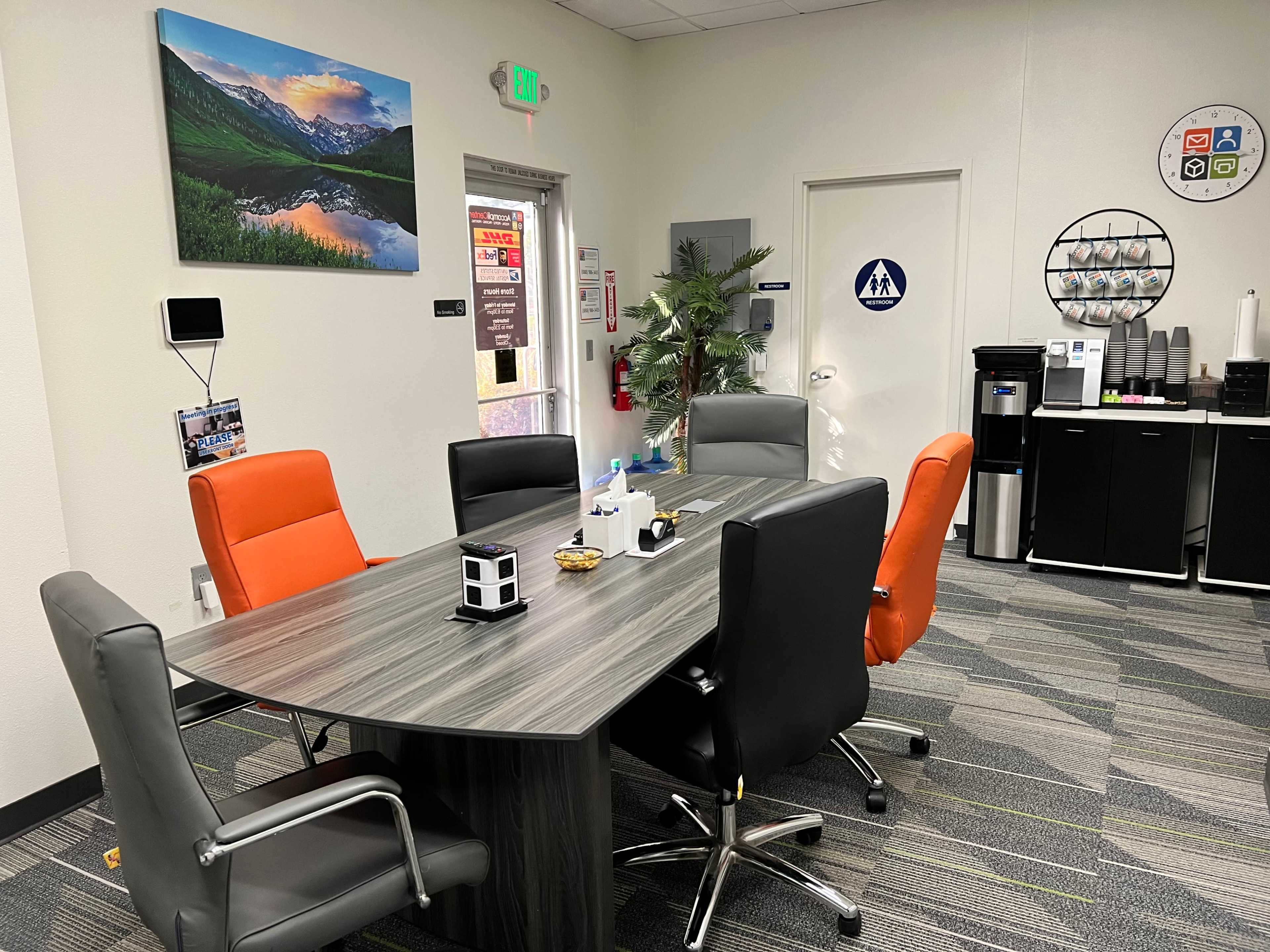 A conference room with a rectangular table surrounded by six chairs, a wall-mounted landscape picture, and a small refreshment area in the corner.
