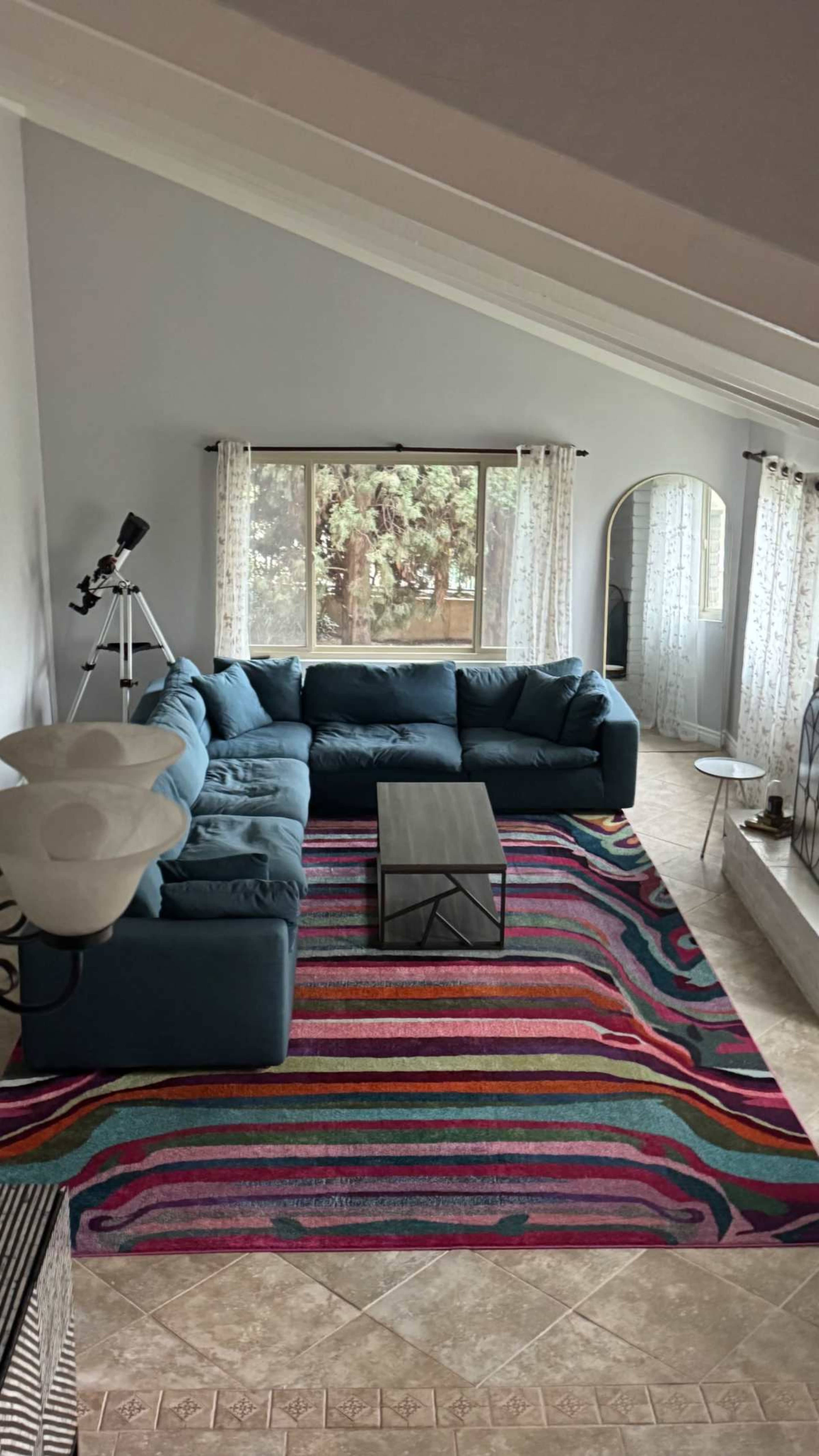 The image shows a spacious living room featuring a large sectional sofa, a coffee table, and a colorful striped area rug, with tall windows allowing natural light to enter.