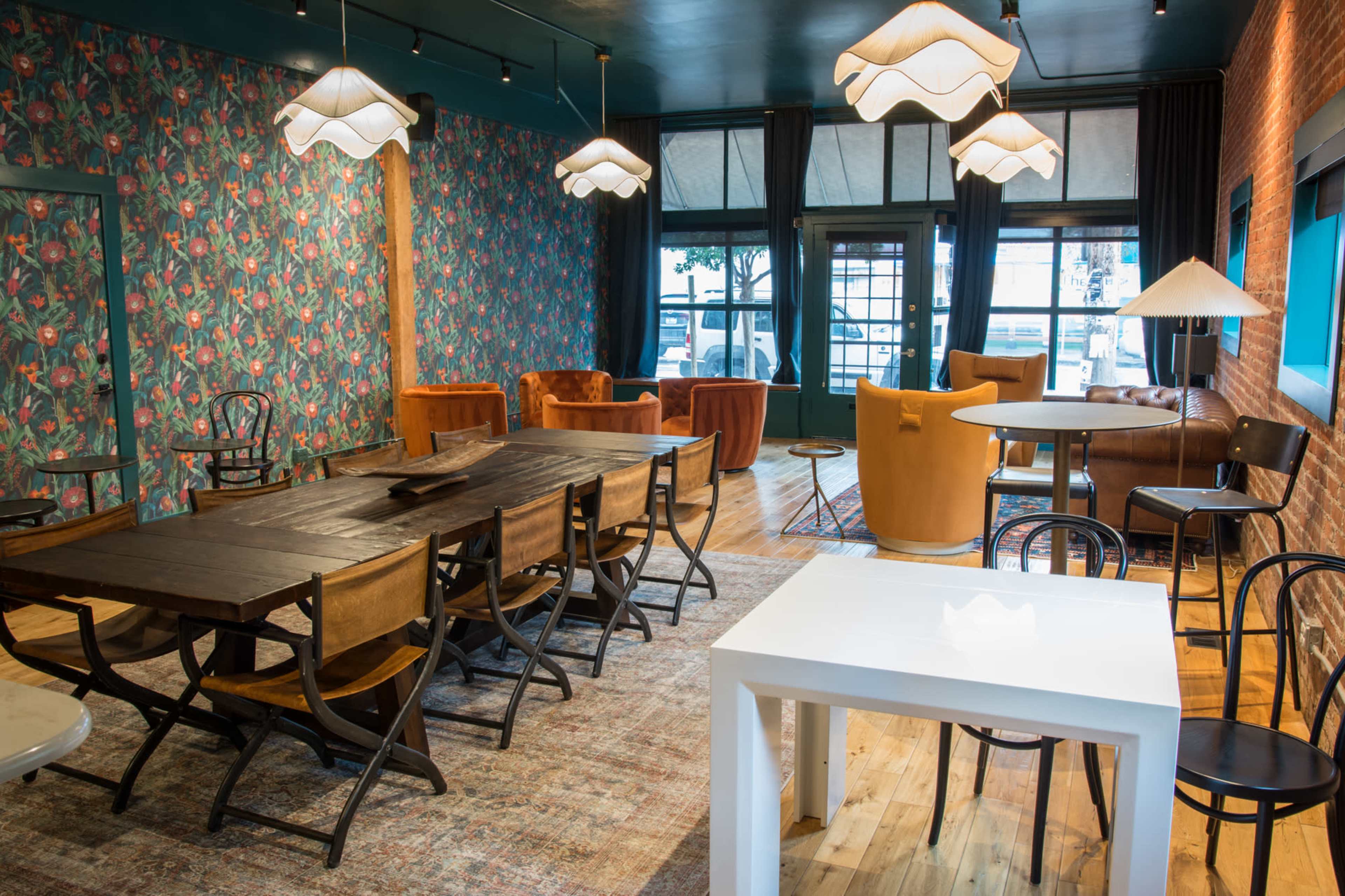 A stylish interior space with patterned wallpaper, a mix of seating options including chairs and sofas, and various dining tables arranged for gatherings.