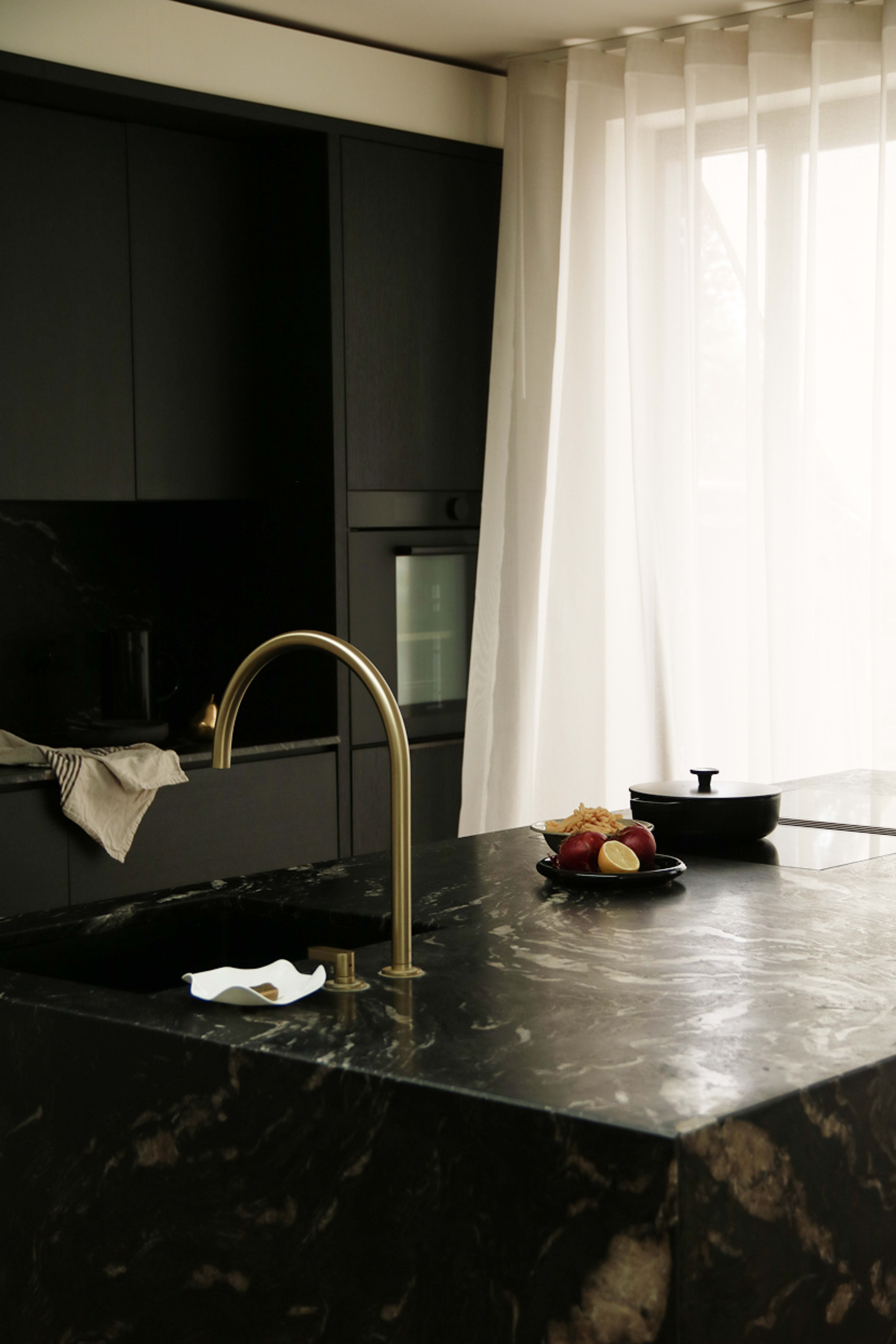 The image shows a modern kitchen with a dark marble island, a gold faucet, and a bowl of apples on a plate next to a black cooking pot.