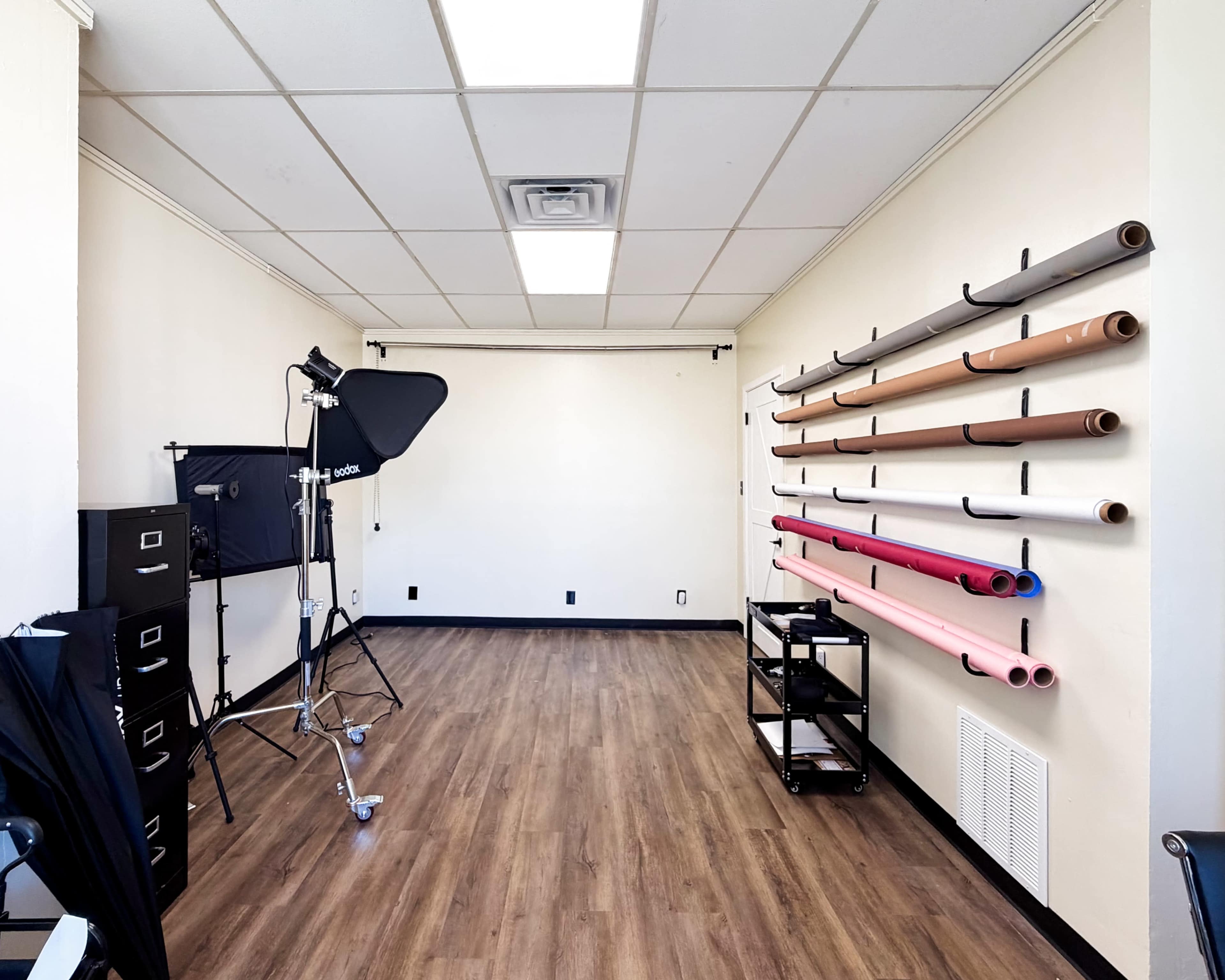 The image shows a photography studio with a backdrop setup, lighting equipment, and paper rolls organized on a wall.
