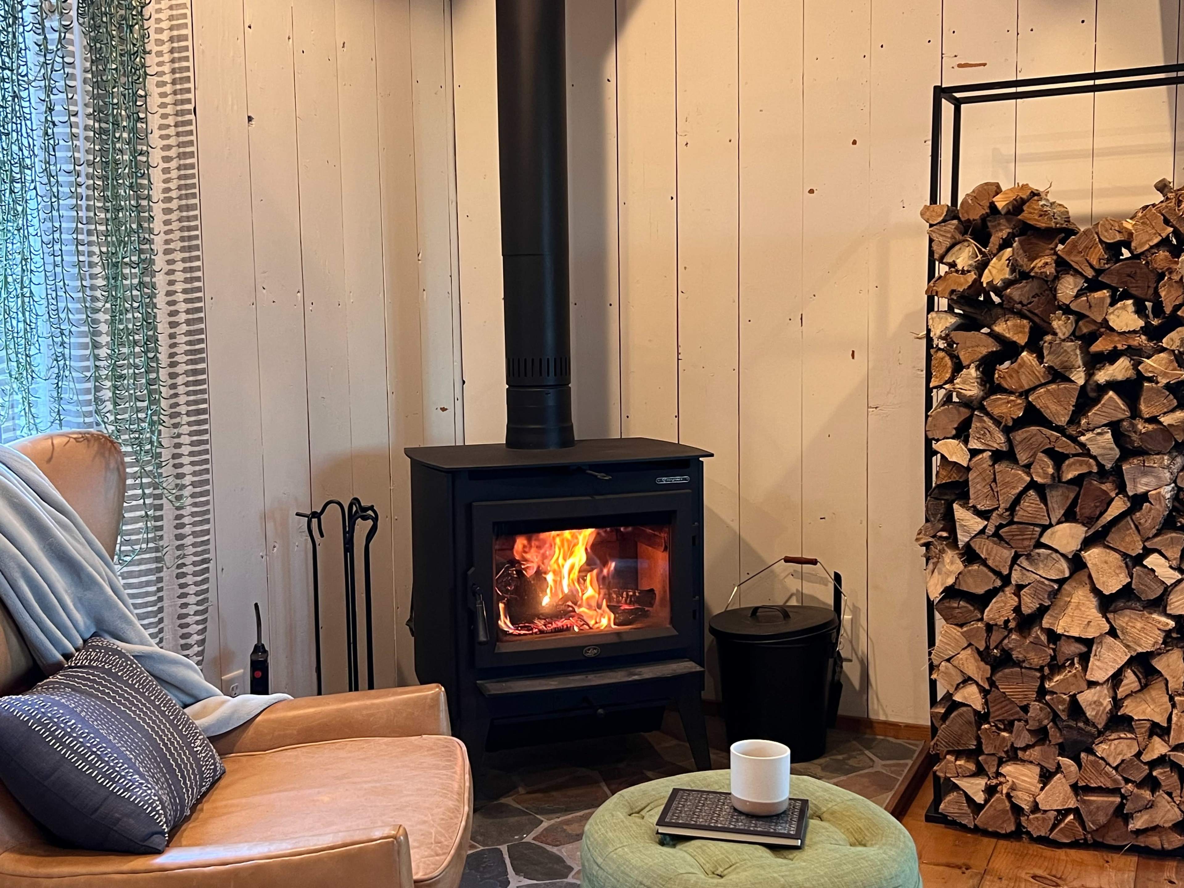 A wood stove with a fire burns beside a stack of firewood in a cozy living space featuring a leather armchair and a small green ottoman.