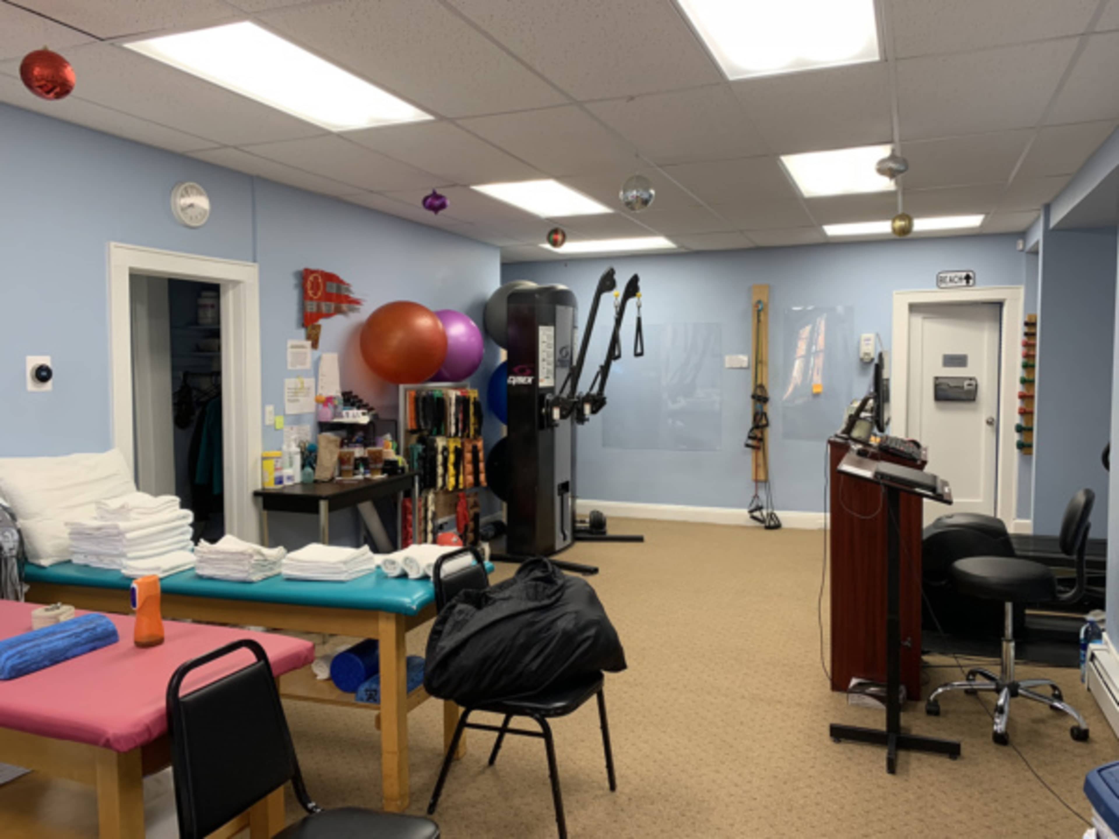 The image shows a fitness or rehabilitation room equipped with exercise equipment, a massage table, and neatly stacked towels.