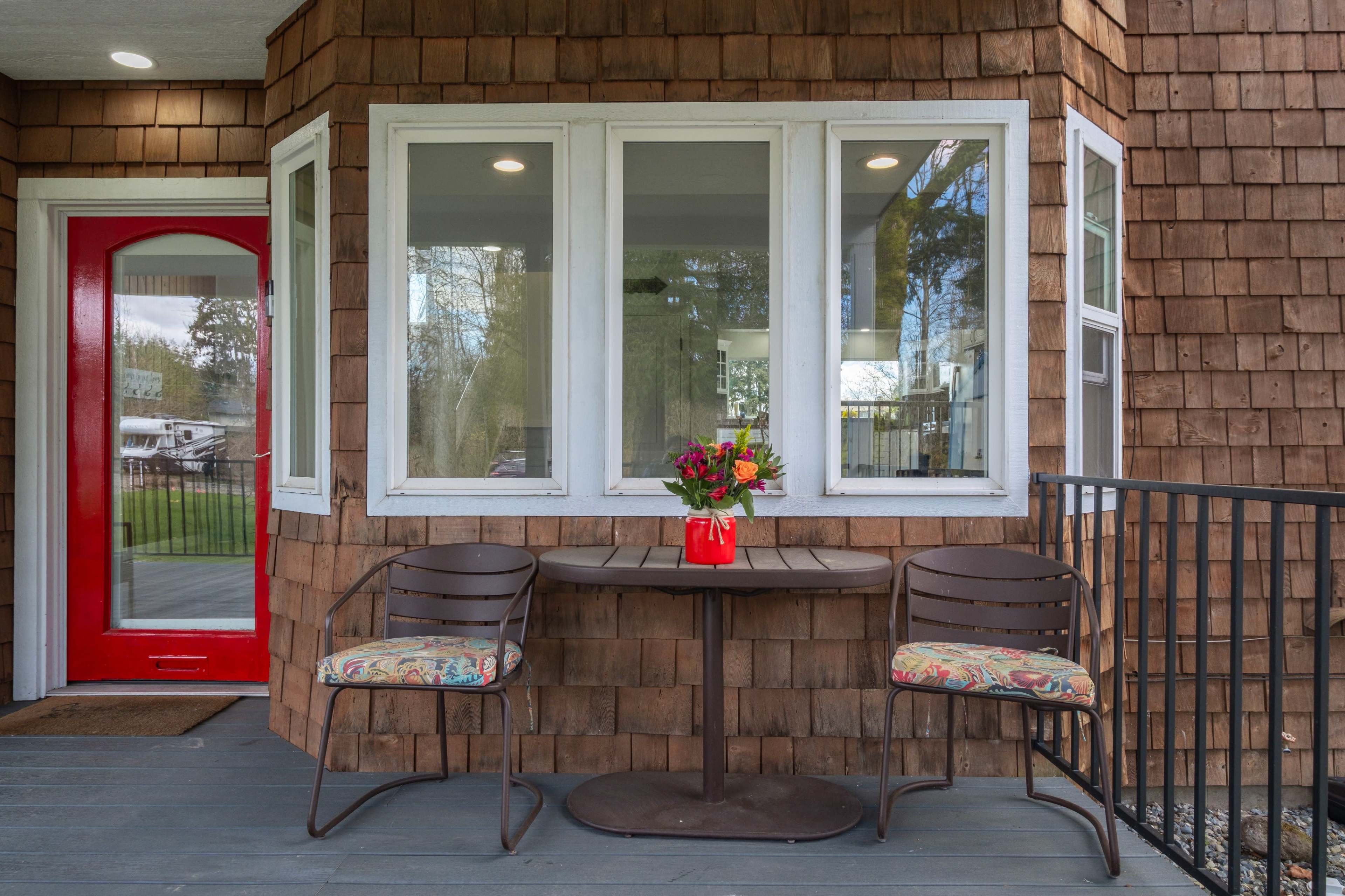 The image shows a corner porch with two chairs and a small round table, adorned with a bouquet of flowers, next to a bright red door.
