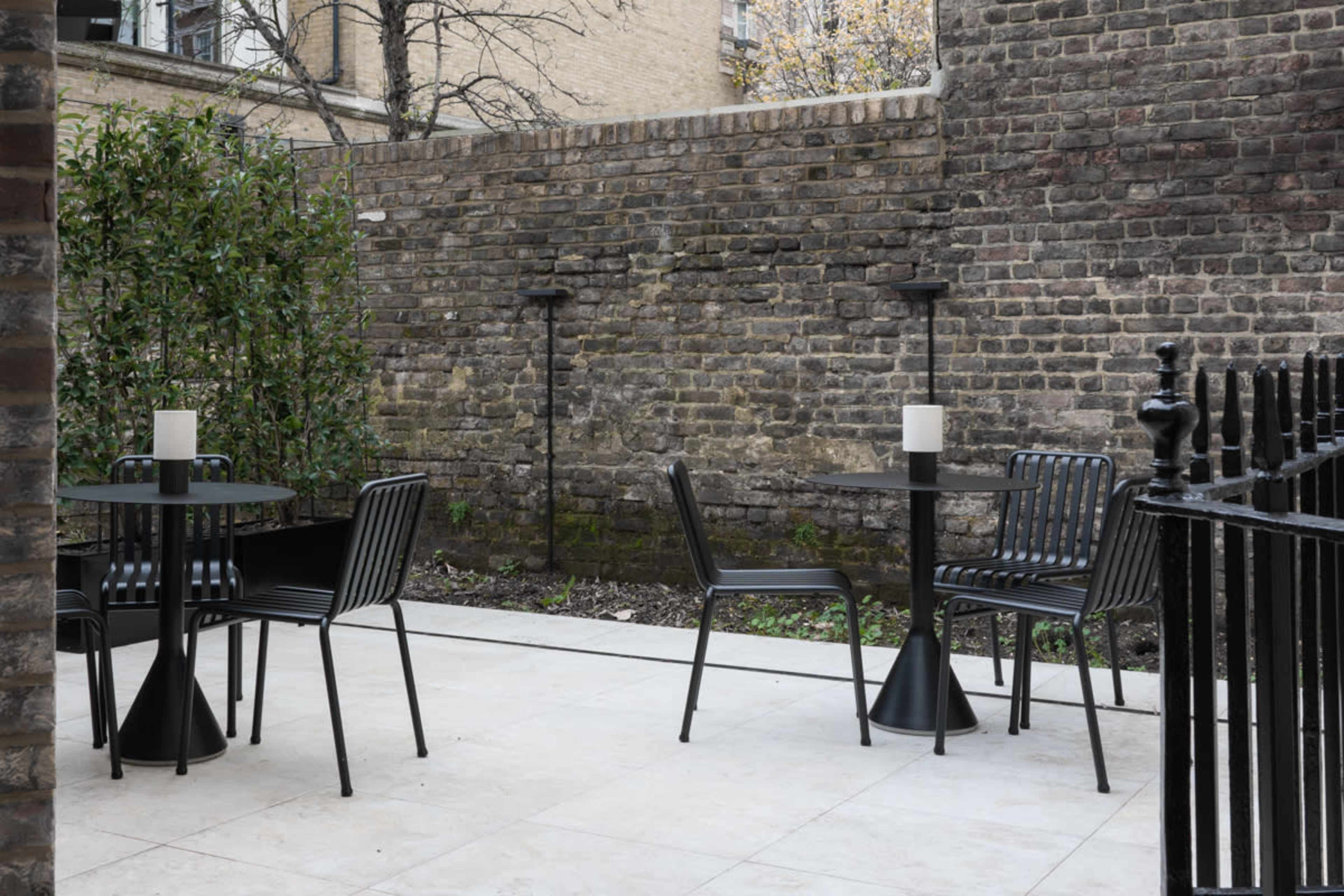 The image shows a small outdoor seating area with two black tables and chairs against a stone wall, surrounded by greenery.
