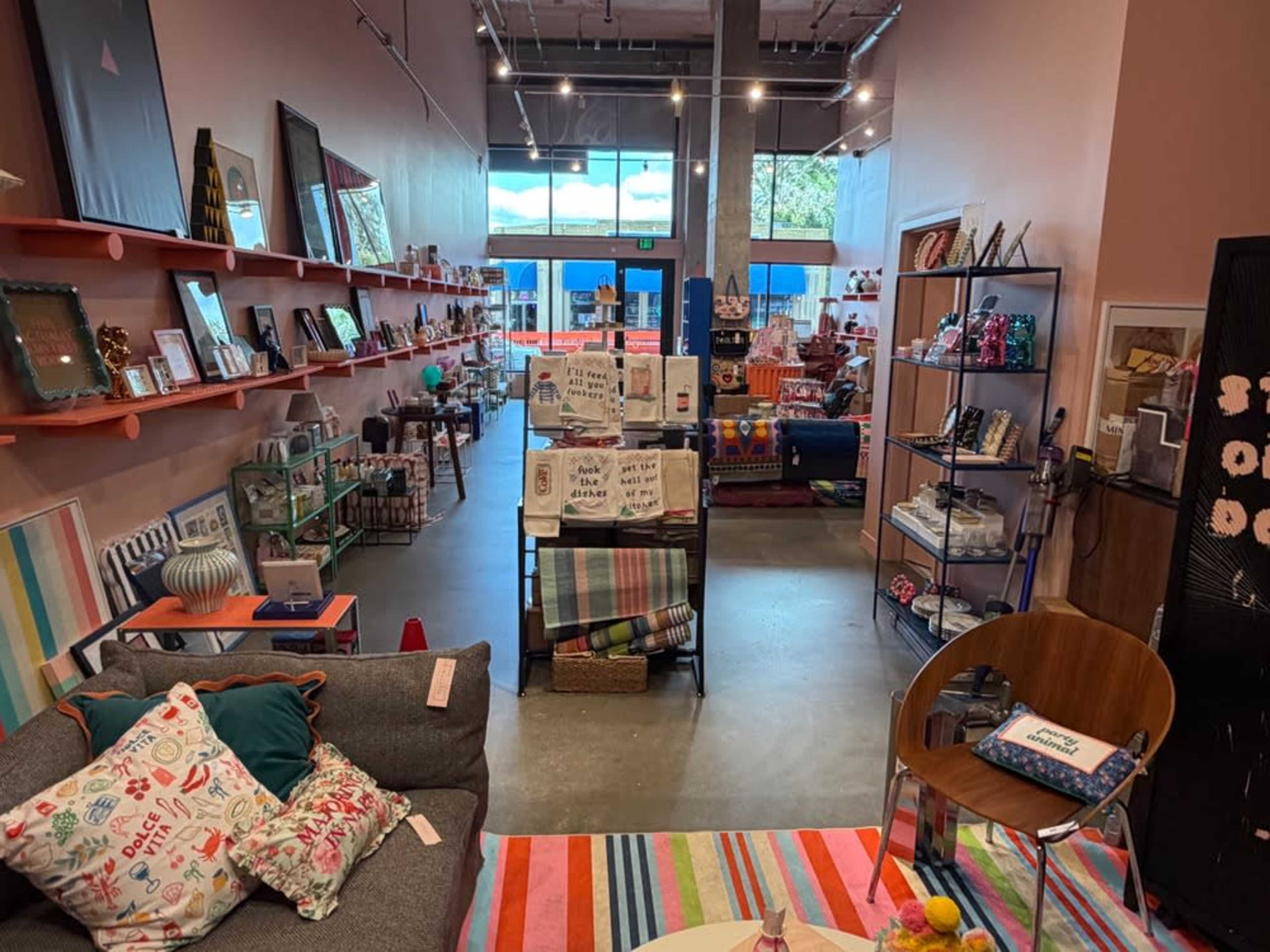 The image shows a brightly lit interior of a boutique filled with various decorative items, furniture, and colorful displays along the walls and floor.