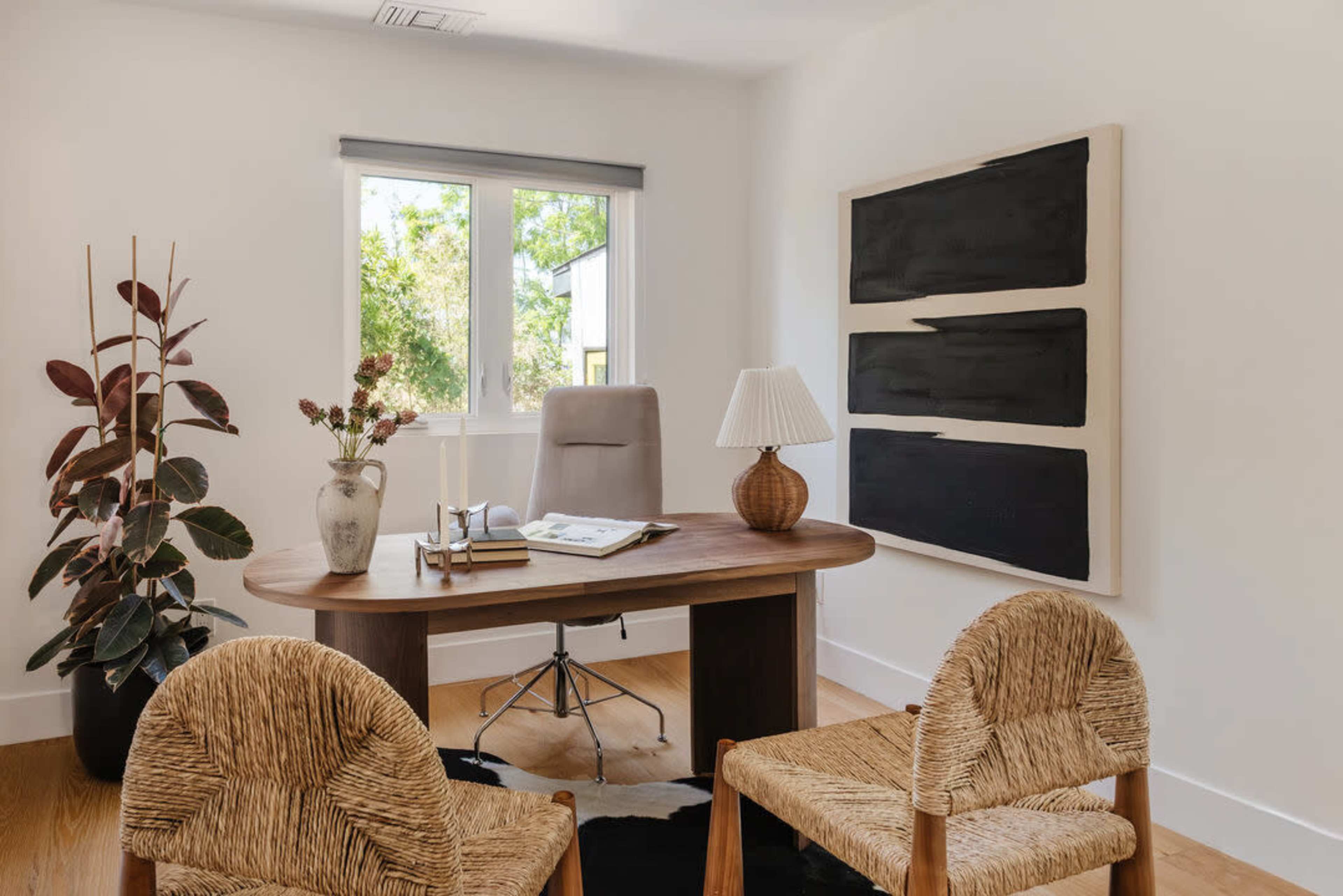 A modern office space features a wooden desk with a chair, two woven chairs, a plant, and an abstract painting on the wall.