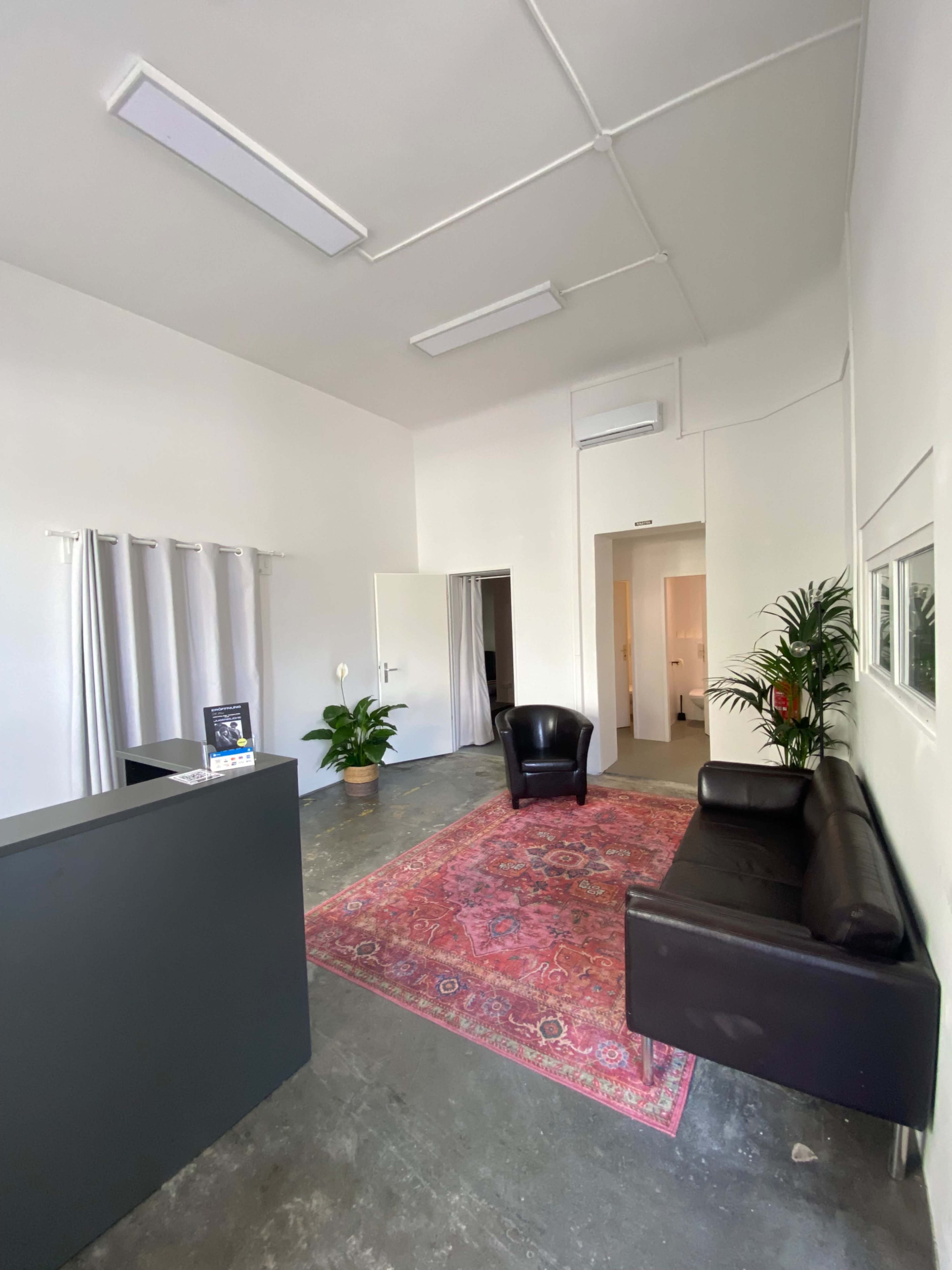 The image shows a minimalist office reception area with a dark reception desk, a black armchair, a leather sofa, a colorful rug, and a potted plant.