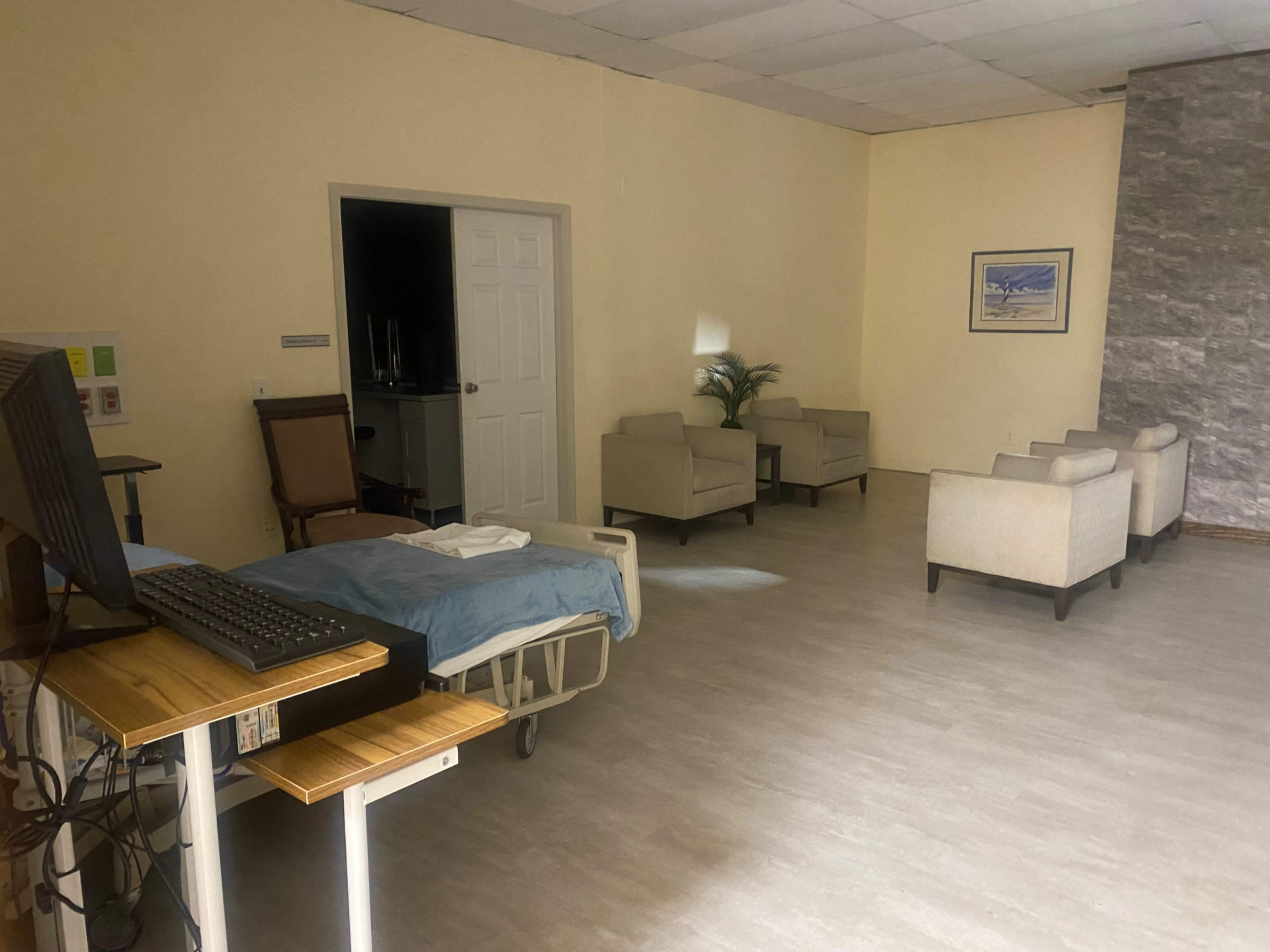 The image shows a hospital room with a bed, a computer on a mobile stand, and several light-colored chairs arranged in a seating area.