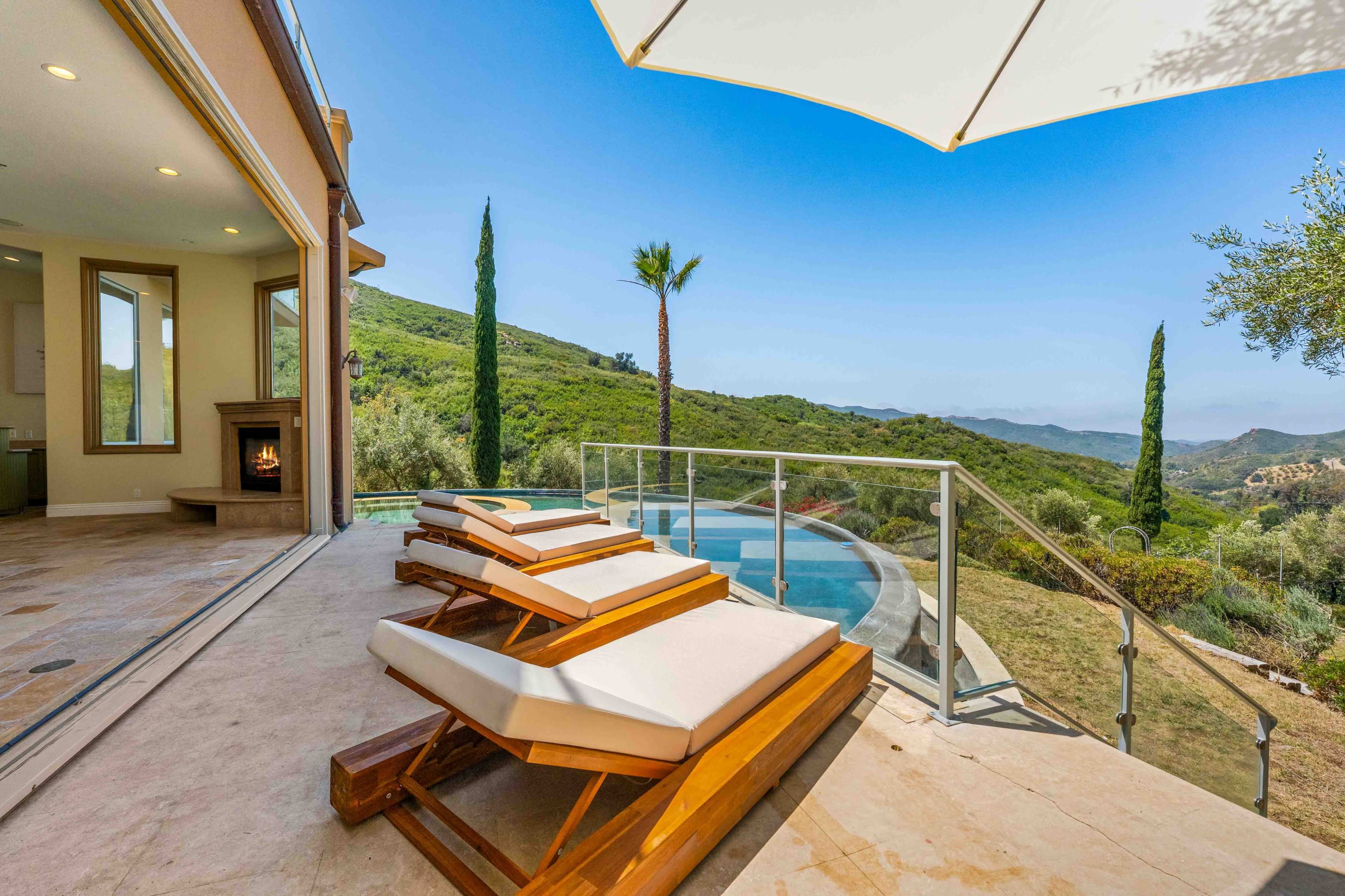 A backyard features lounge chairs by a swimming pool, with a scenic view of rolling hills and palm trees in the background.
