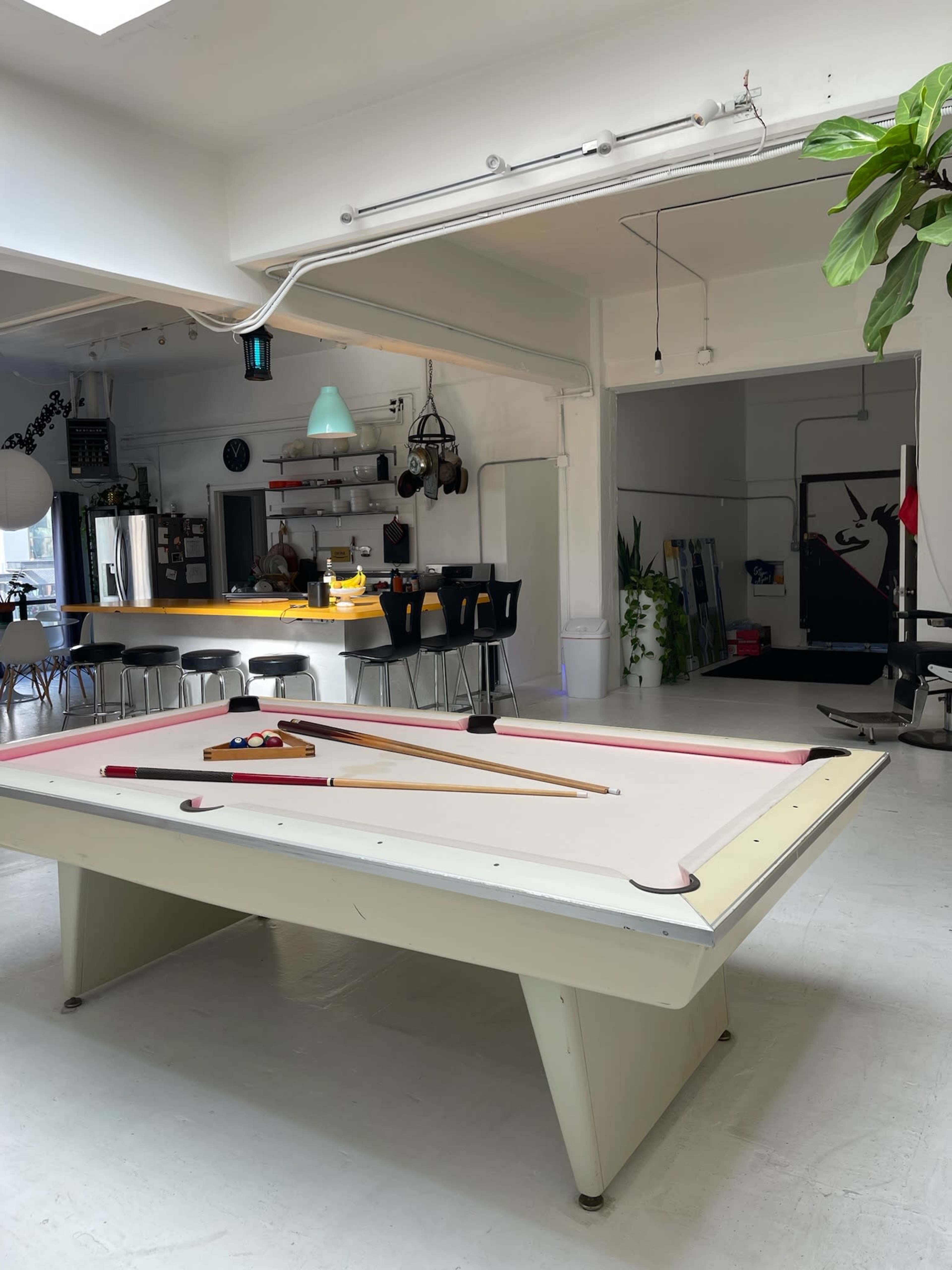 The image shows a bright, open interior space featuring a pool table in the foreground, with a kitchen area and seating visible in the background.
