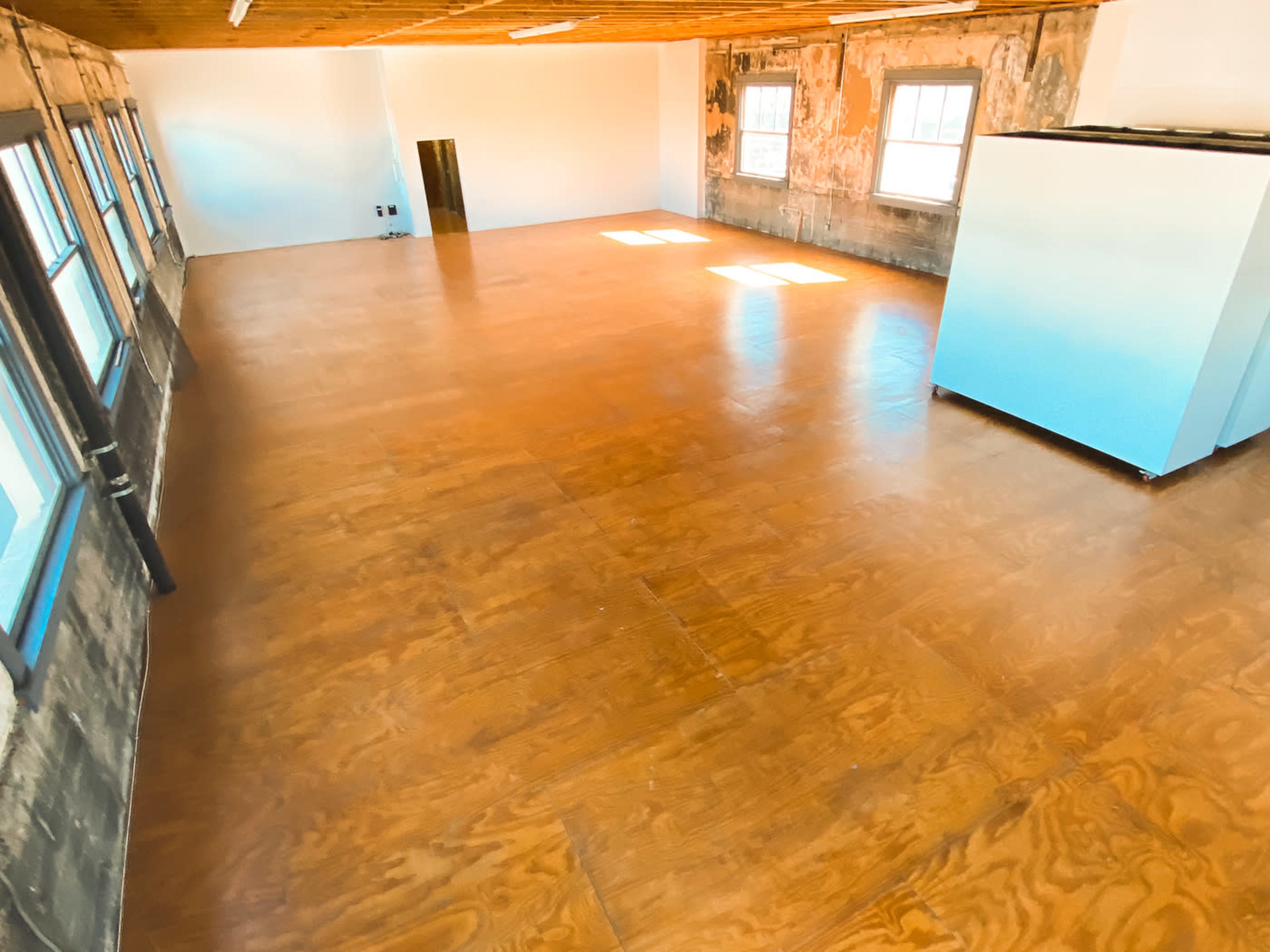 The image shows an empty room with wooden flooring, exposed brick walls, and large windows allowing light to enter, with a white appliance visible in one corner.