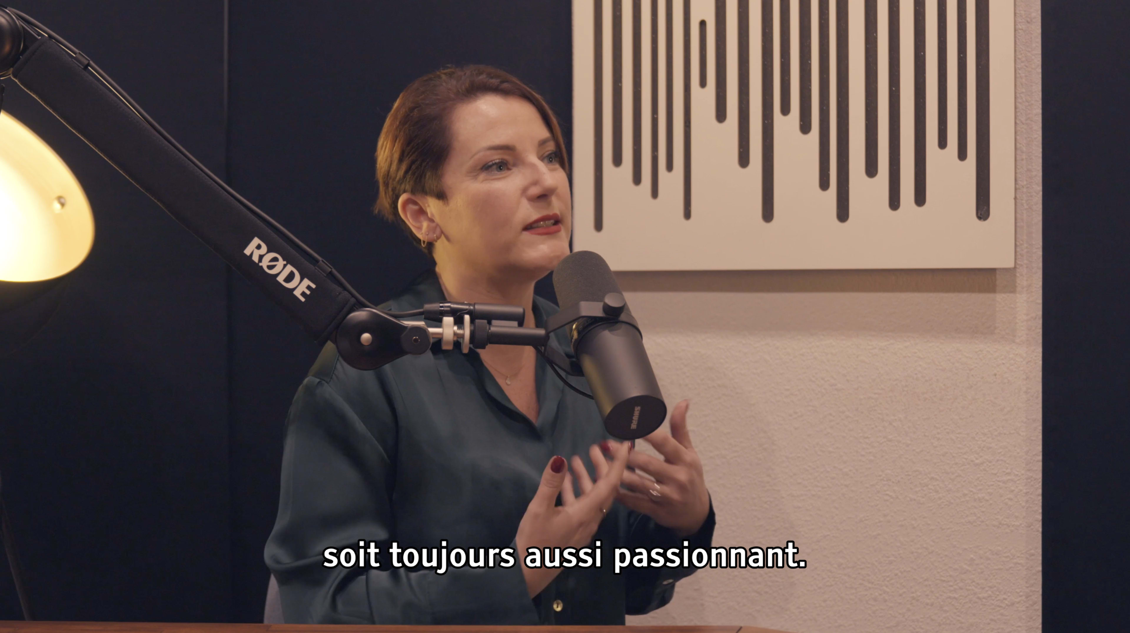 A woman speaks into a microphone at a desk while gesturing, with a modern graphic design on the wall behind her.