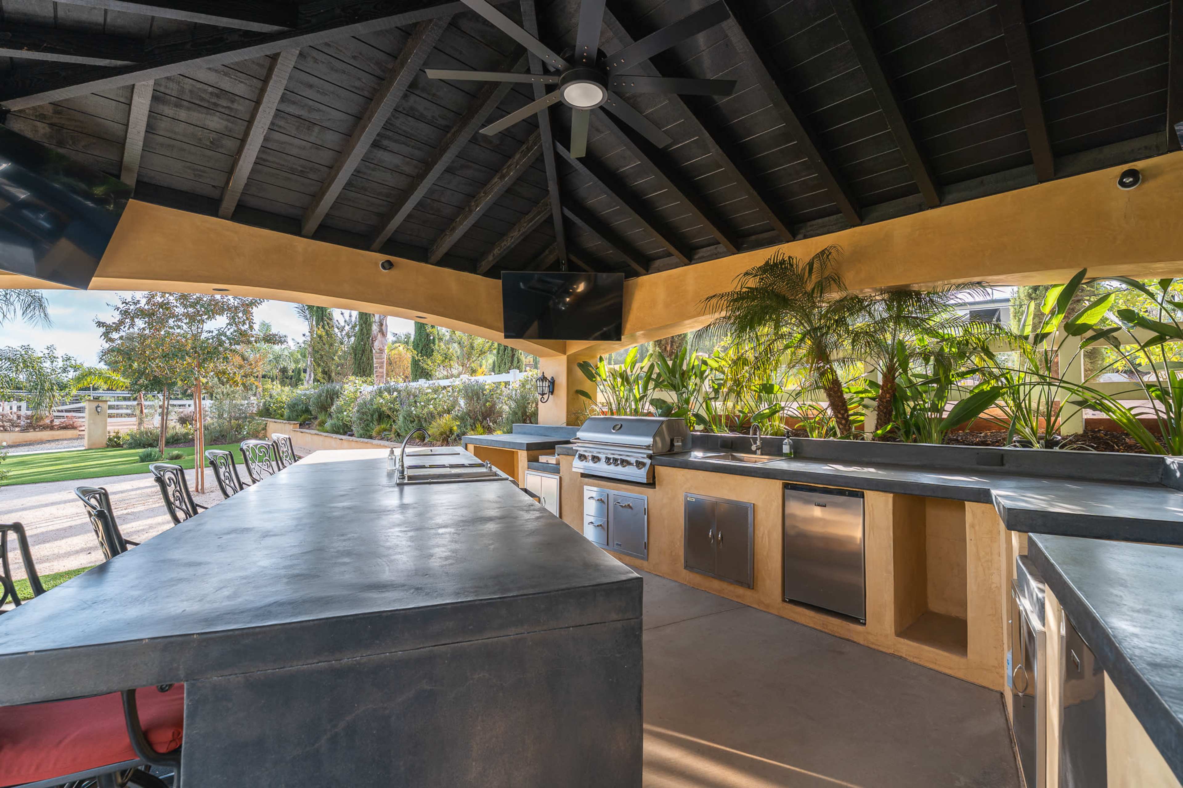 The image shows a spacious outdoor kitchen under a shaded structure with a black wooden ceiling, featuring a stainless steel grill and countertop, surrounded by lush greenery.