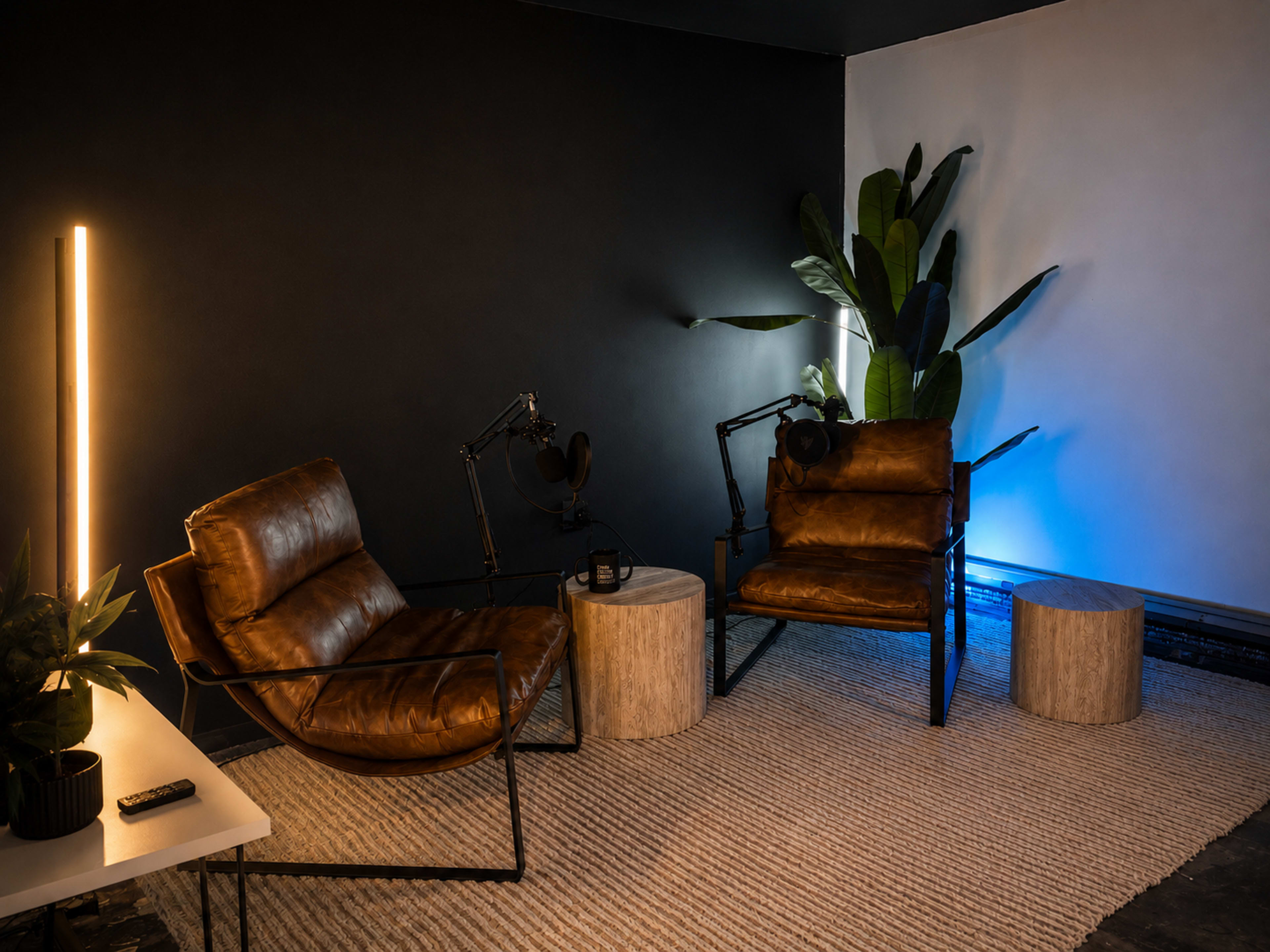 The image shows a modern interior corner featuring two brown leather chairs, a circular wooden side table, and a tall plant, with contrasting lighting from a floor lamp and ambient blue lights.