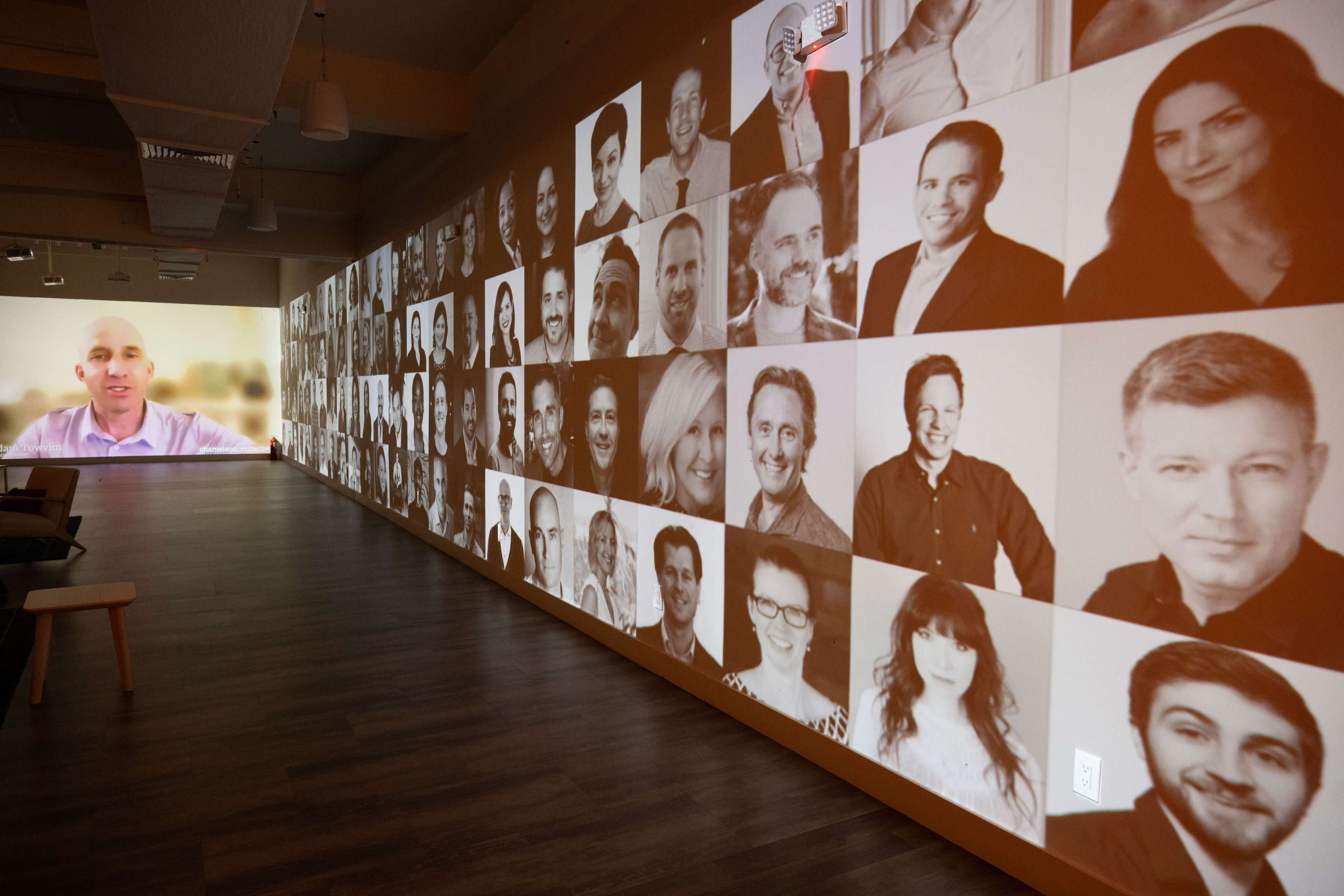 A wall displays a collage of monochrome headshots of various people, while a separate projection shows a video of a speaker in a purple shirt.