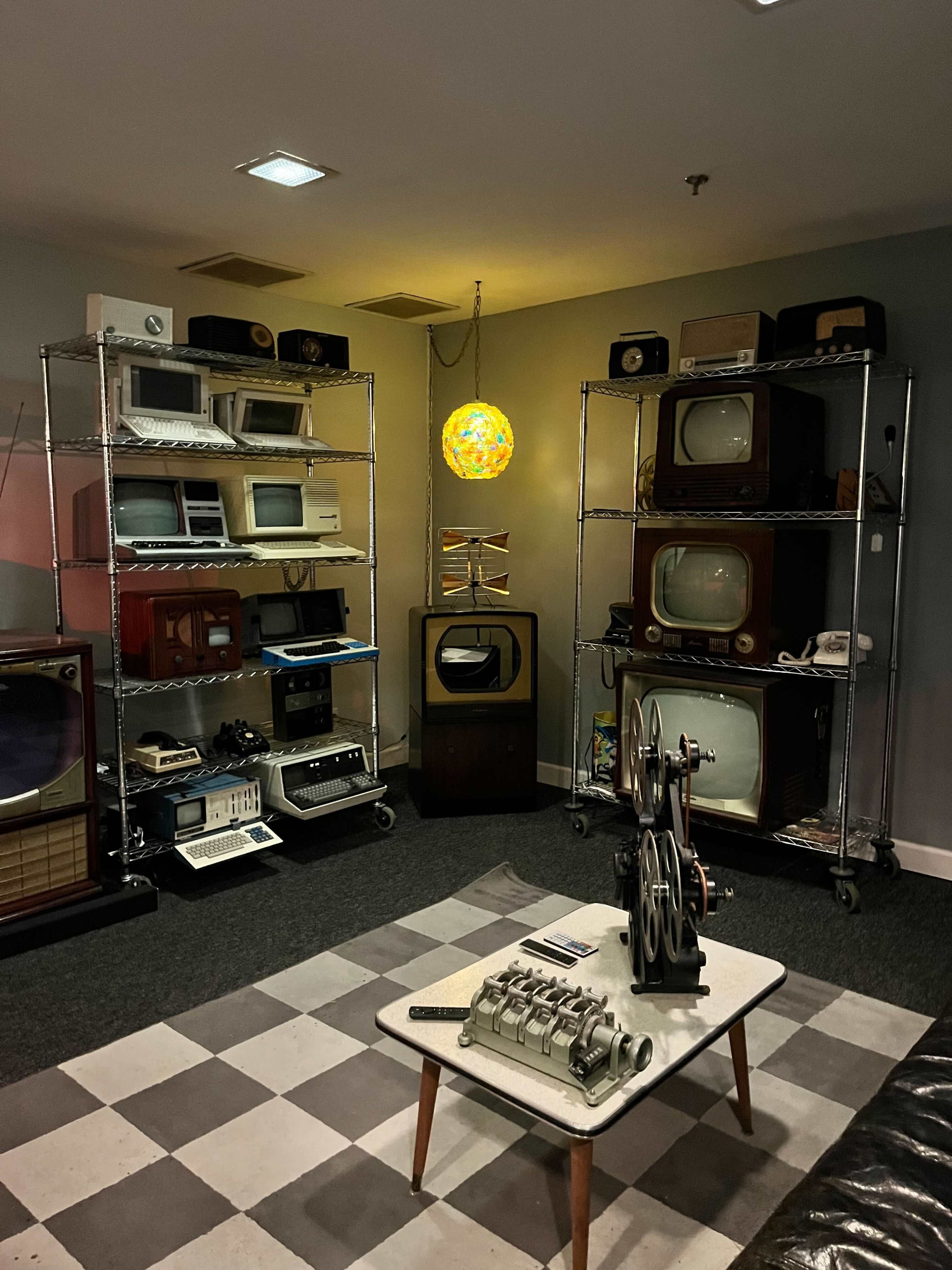 The image features a room displaying an assortment of vintage televisions and electronic devices, arranged on metal shelving units, with a retro lamp illuminating the space.