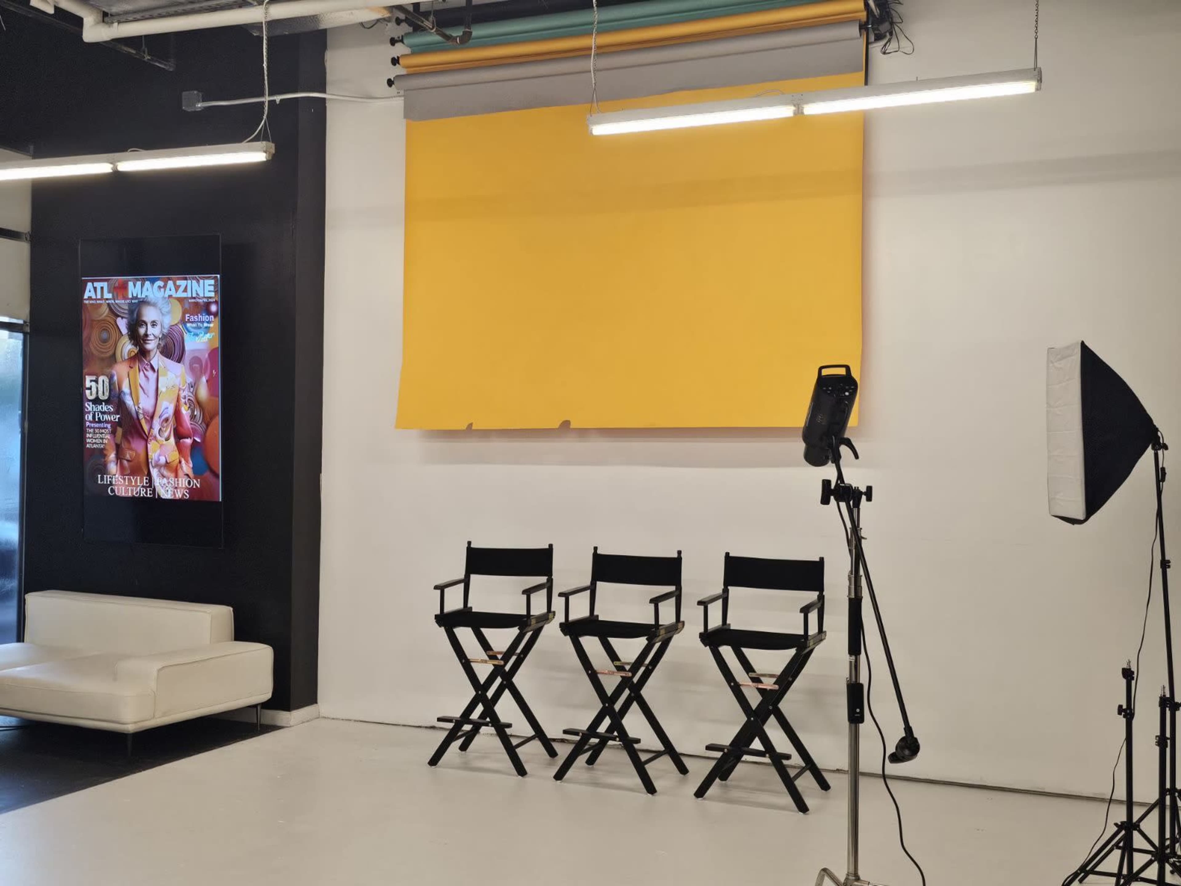 The image shows a studio setup featuring three black director's chairs in front of a yellow backdrop and a lit light stand, along with a magazine cover displayed on the wall.