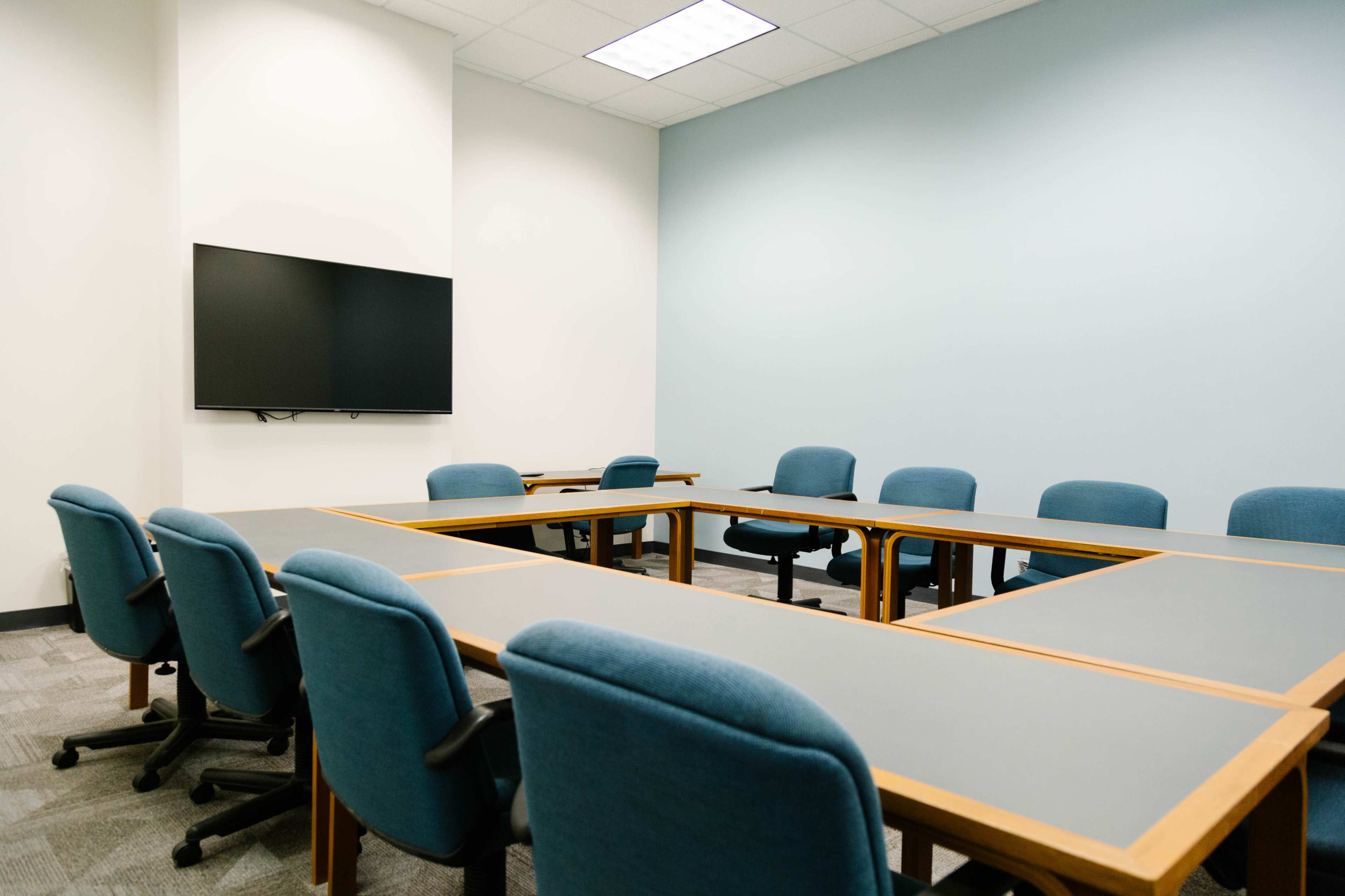 A conference room features a rectangular arrangement of tables surrounded by blue chairs and a wall-mounted television.