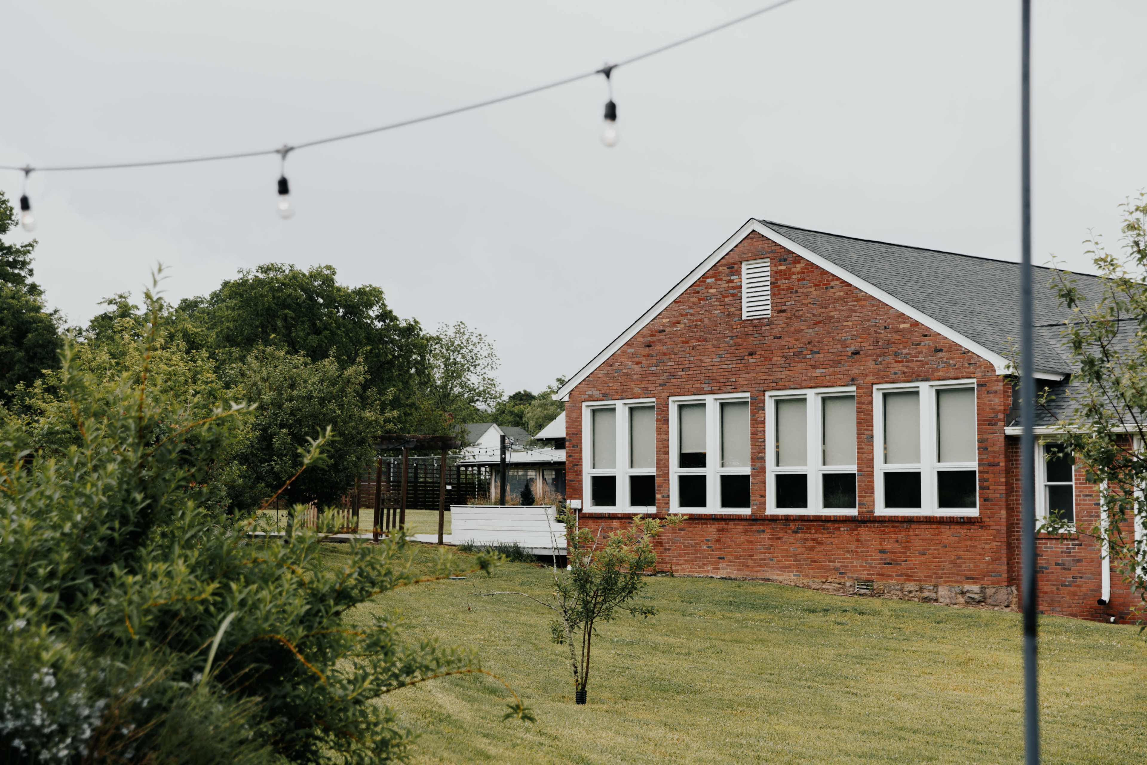 A brick house with multiple large windows is set against a backdrop of green trees and grass, with string lights hanging in the foreground.