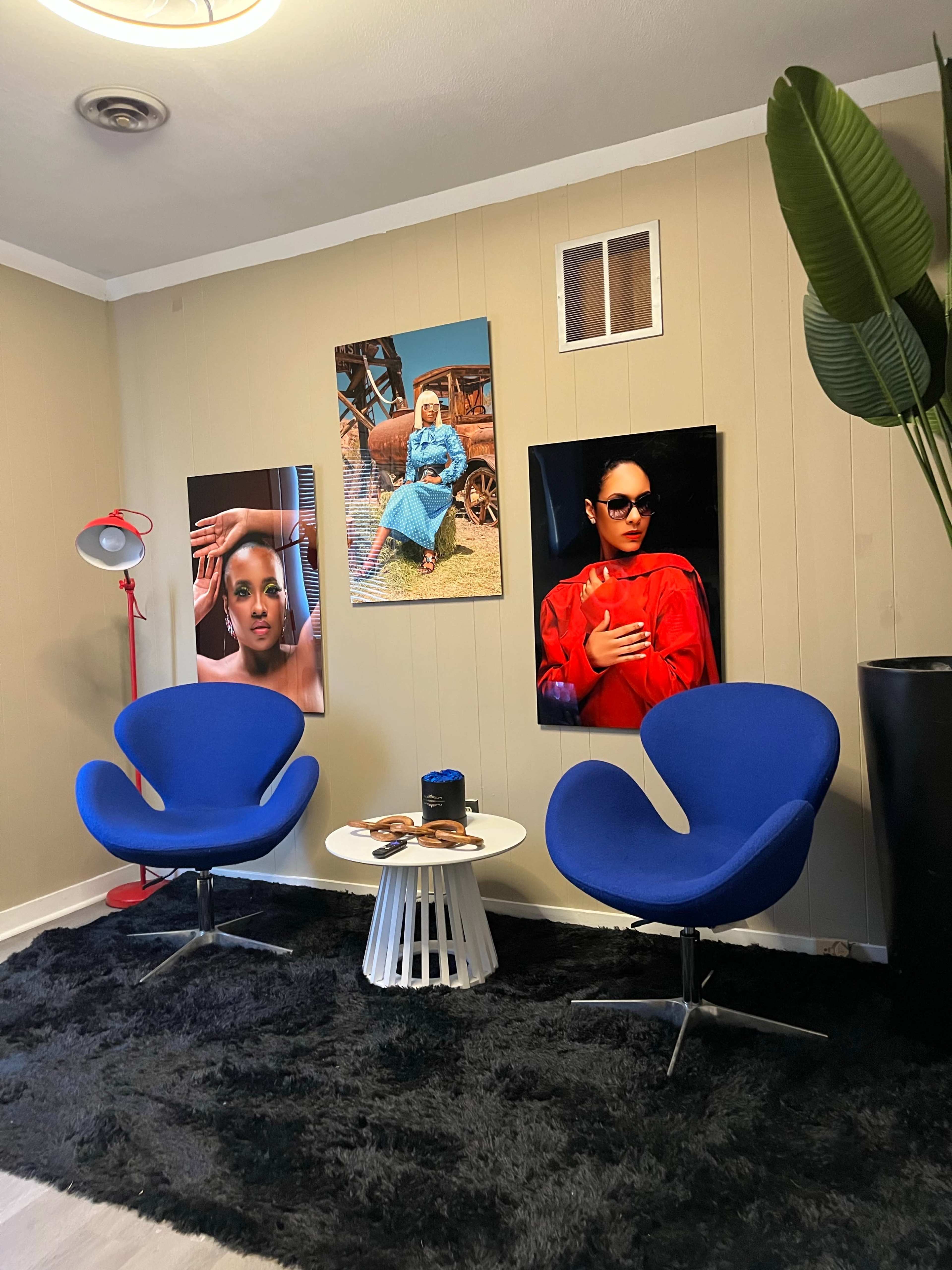 The image shows a modern living space with two blue chairs, a round table, and two large portraits on the walls, accompanied by a potted plant and a lamp.