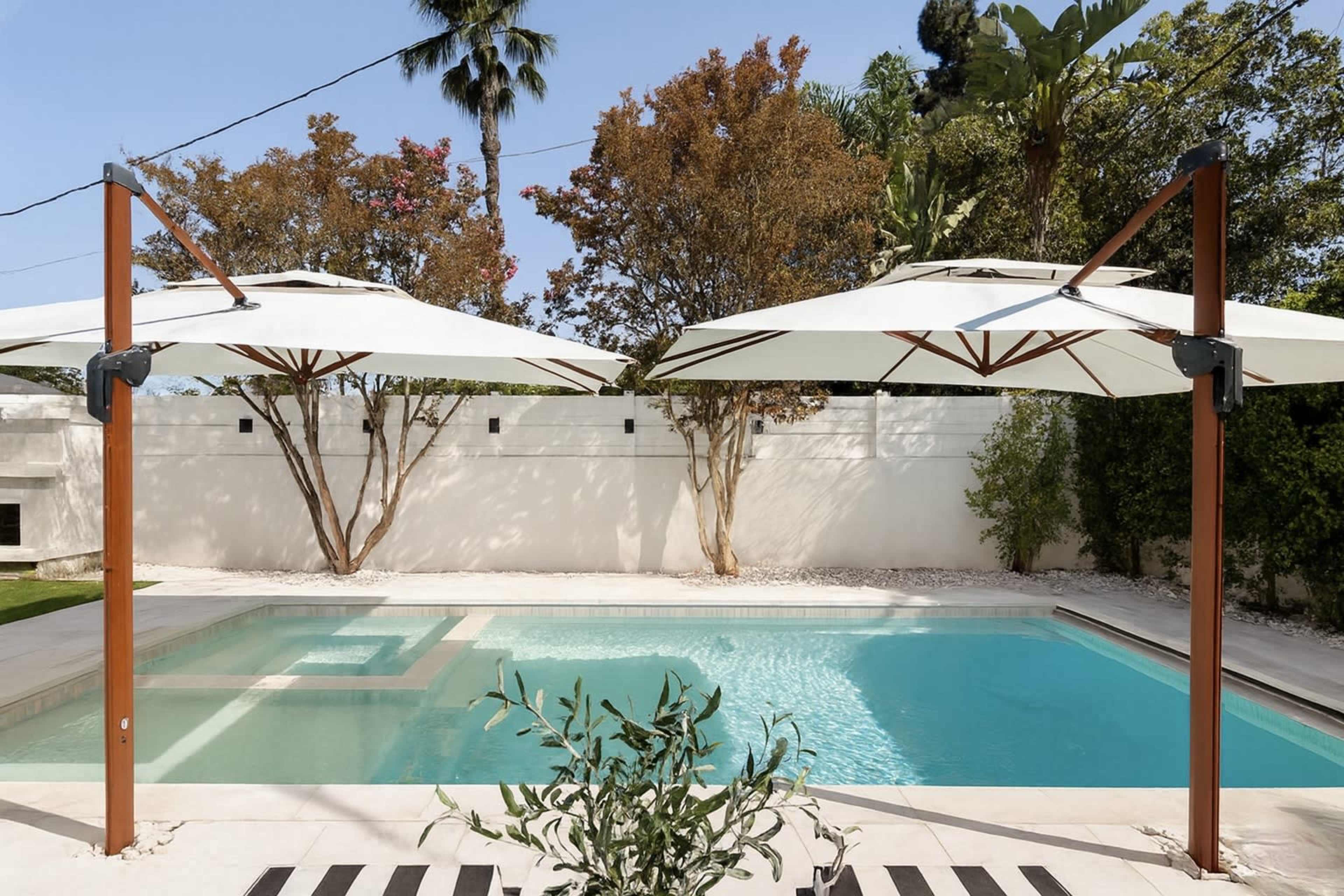 The image shows a swimming pool surrounded by white umbrellas and trees in a landscaped outdoor area.