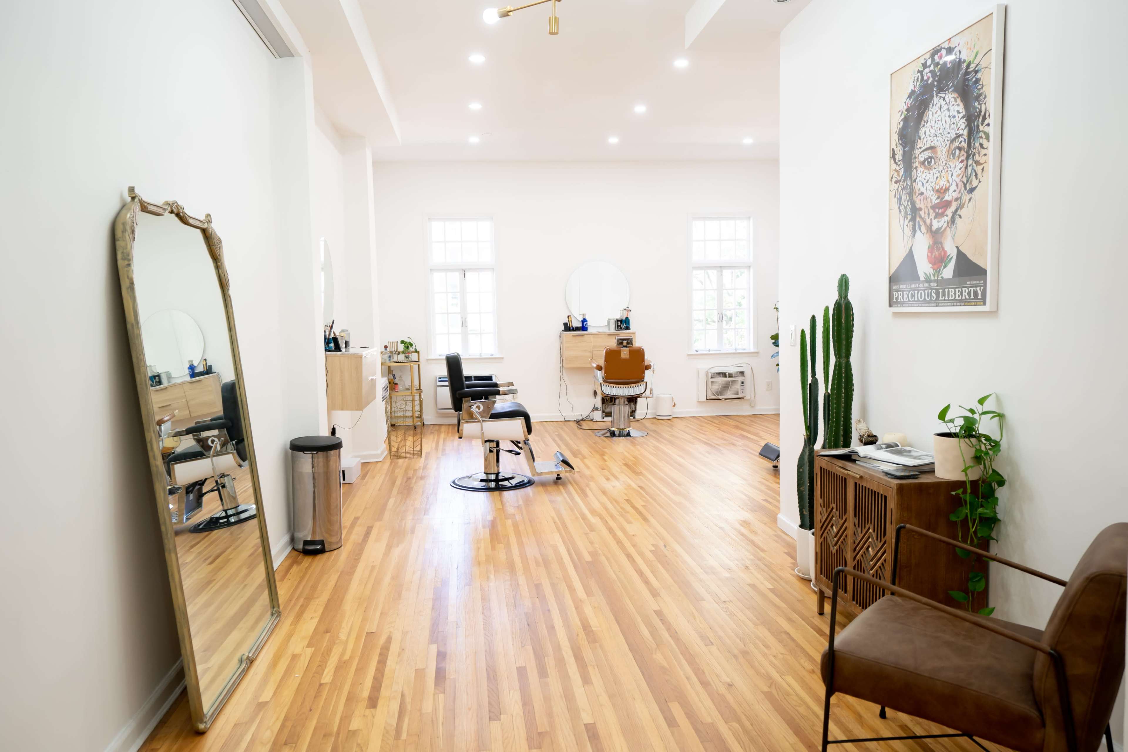 A well-lit salon features wooden flooring, a large mirror, hair styling stations, and green plants.