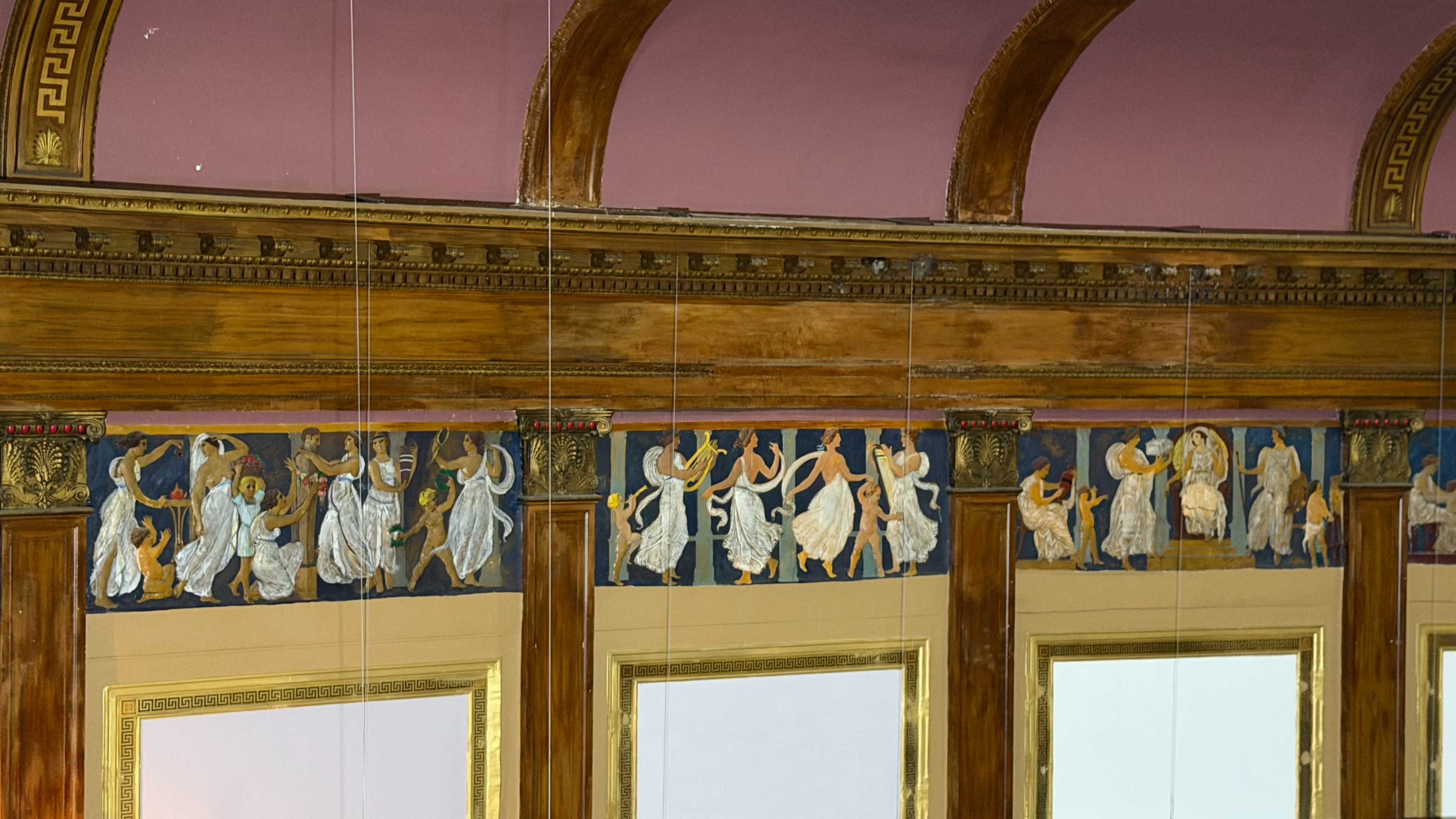 The image shows a detailed mural depicting figures in flowing robes engaged in various dances and poses along a wall beneath a wooden decorative border.
