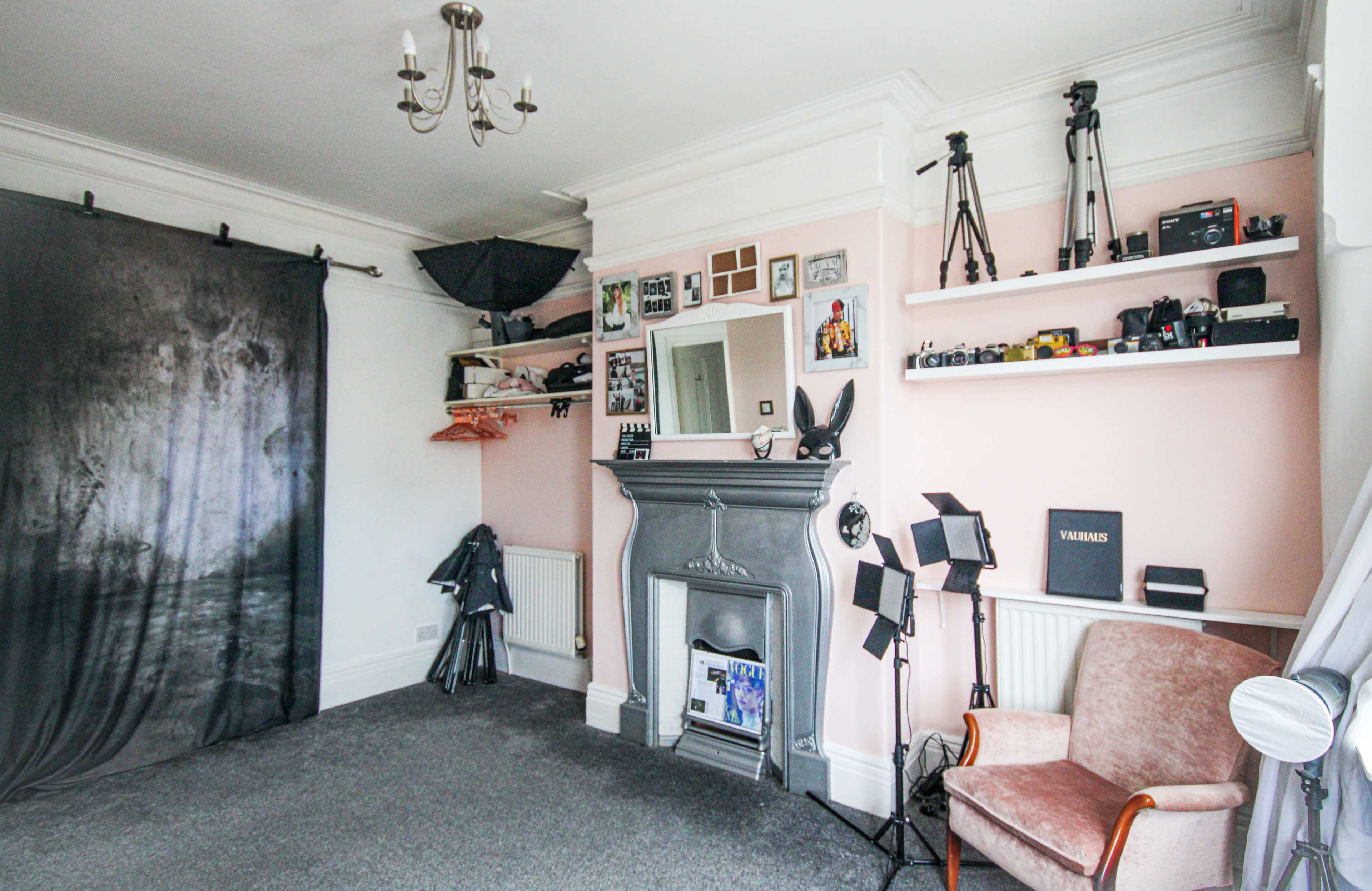 The image shows a room with a grey carpet, a pink wall featuring shelves with cameras, and a fireplace flanked by a chair and photography equipment.