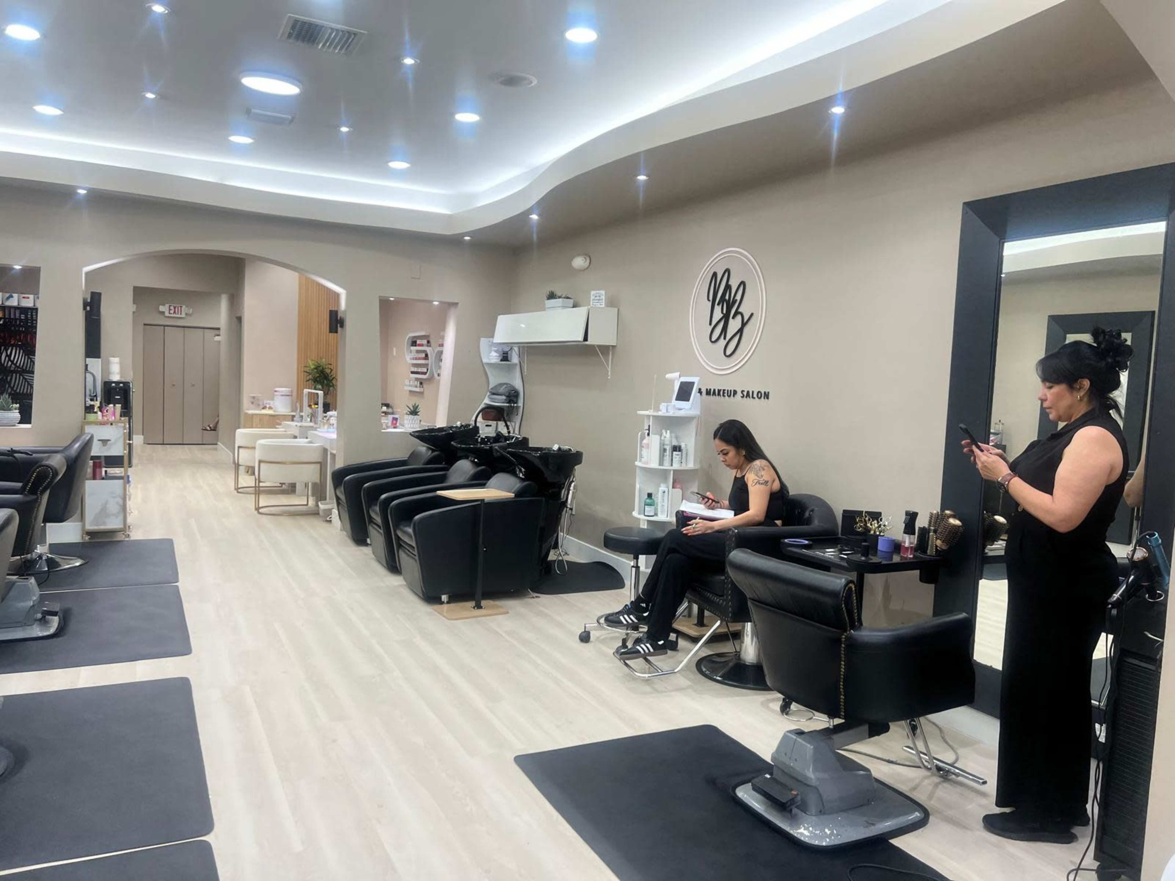 The image shows the interior of a modern makeup salon with several styling chairs, mirrors, and a waiting area where two women are engaged in activities on their phones and reading.