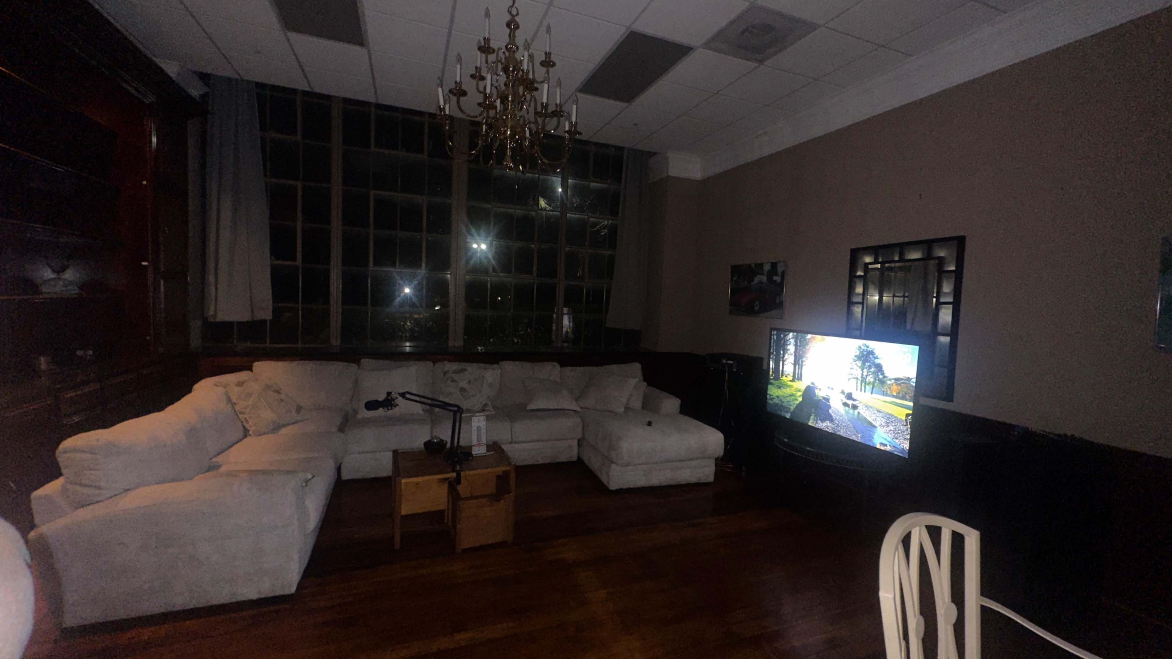 The image shows a living room with a large sectional couch, a coffee table, a television displaying a scenic image, and a chandelier overhead.