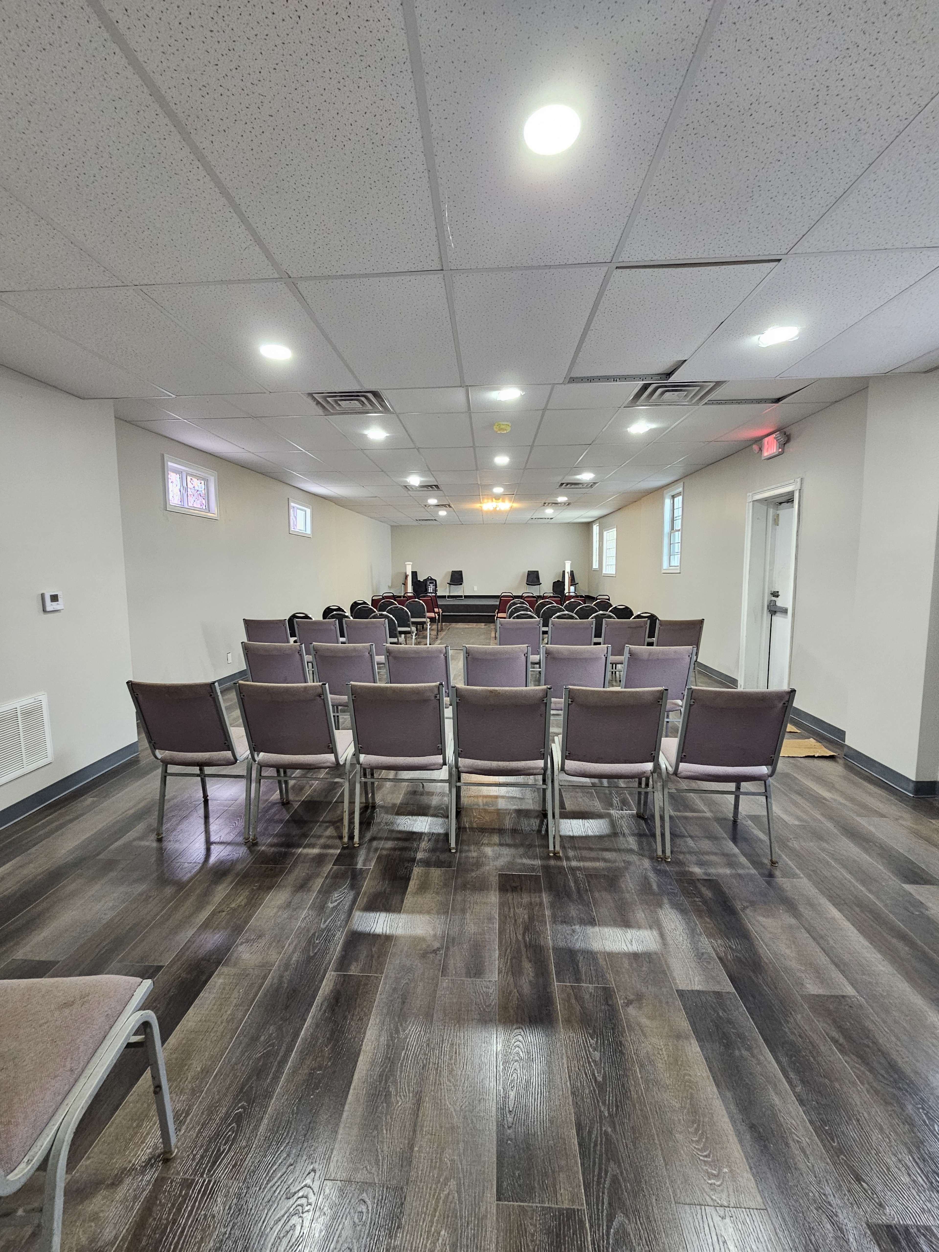 The image shows a well-lit interior room set up with rows of gray chairs facing a stage or focal point, featuring wooden floors and a few windows along the walls.