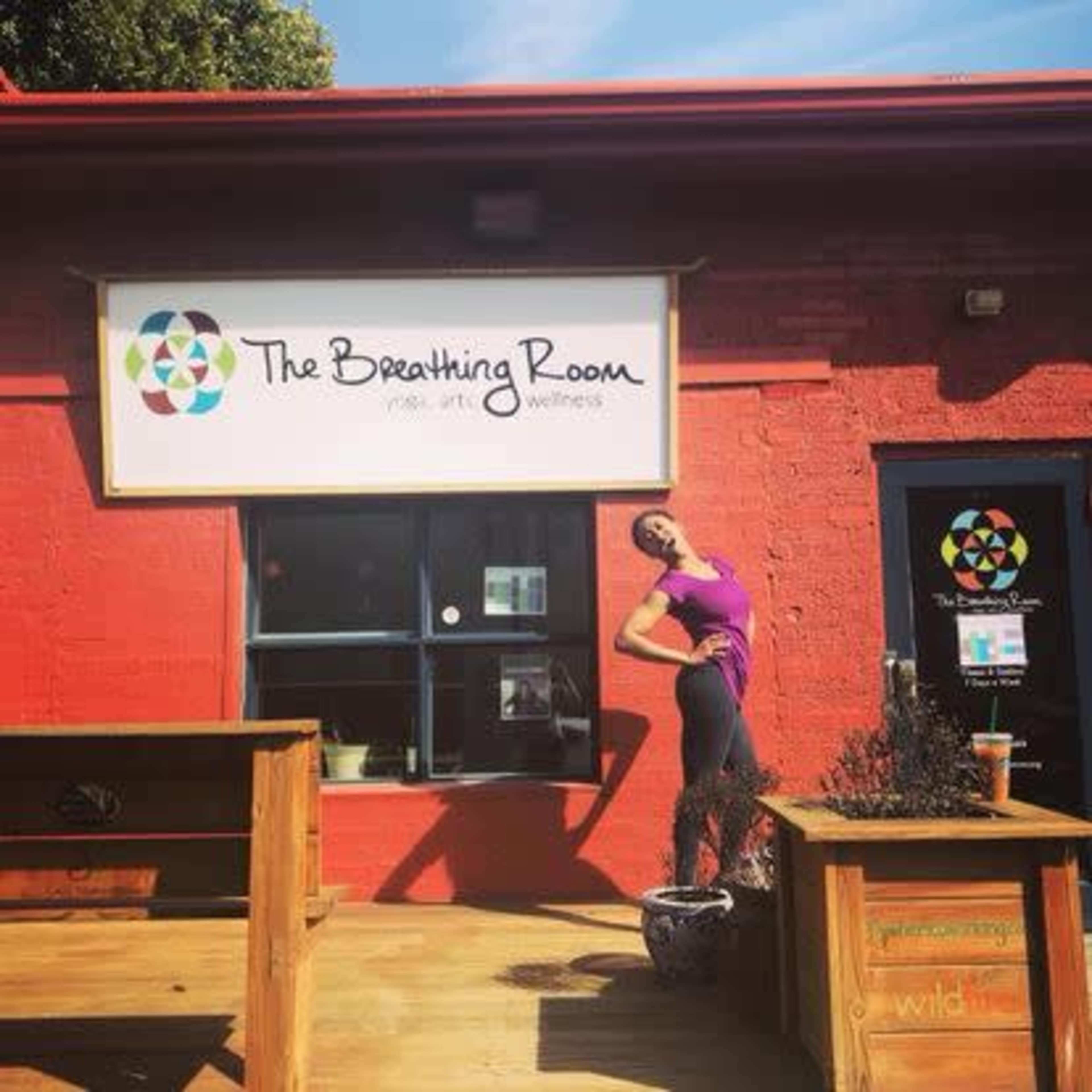 A woman poses in front of a vibrant red building with a sign reading "The Breathing Room" and a decorative logo.