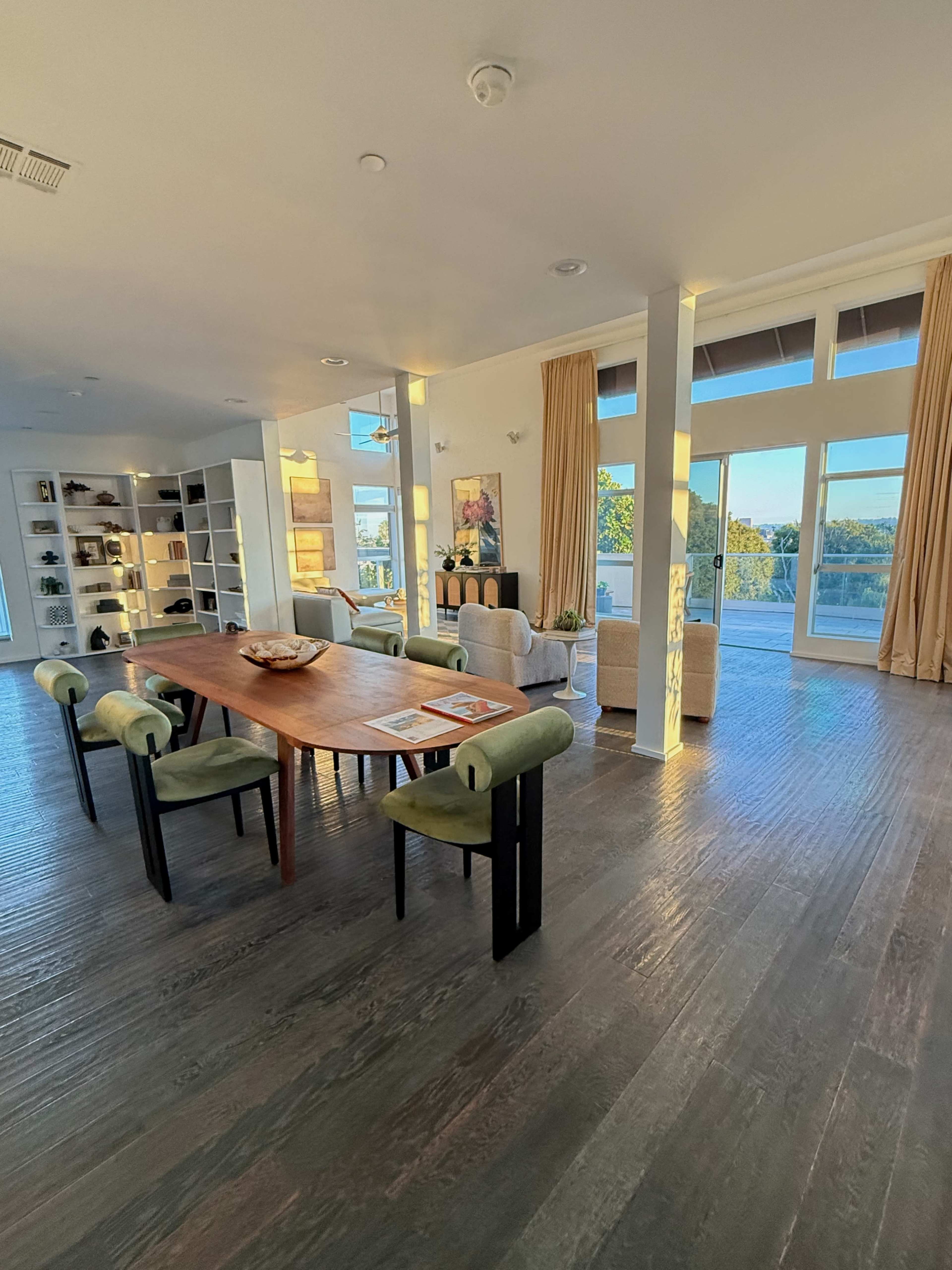 The image shows a spacious, contemporary dining area with a wooden table, upholstered chairs, and large windows offering views of the outdoors.