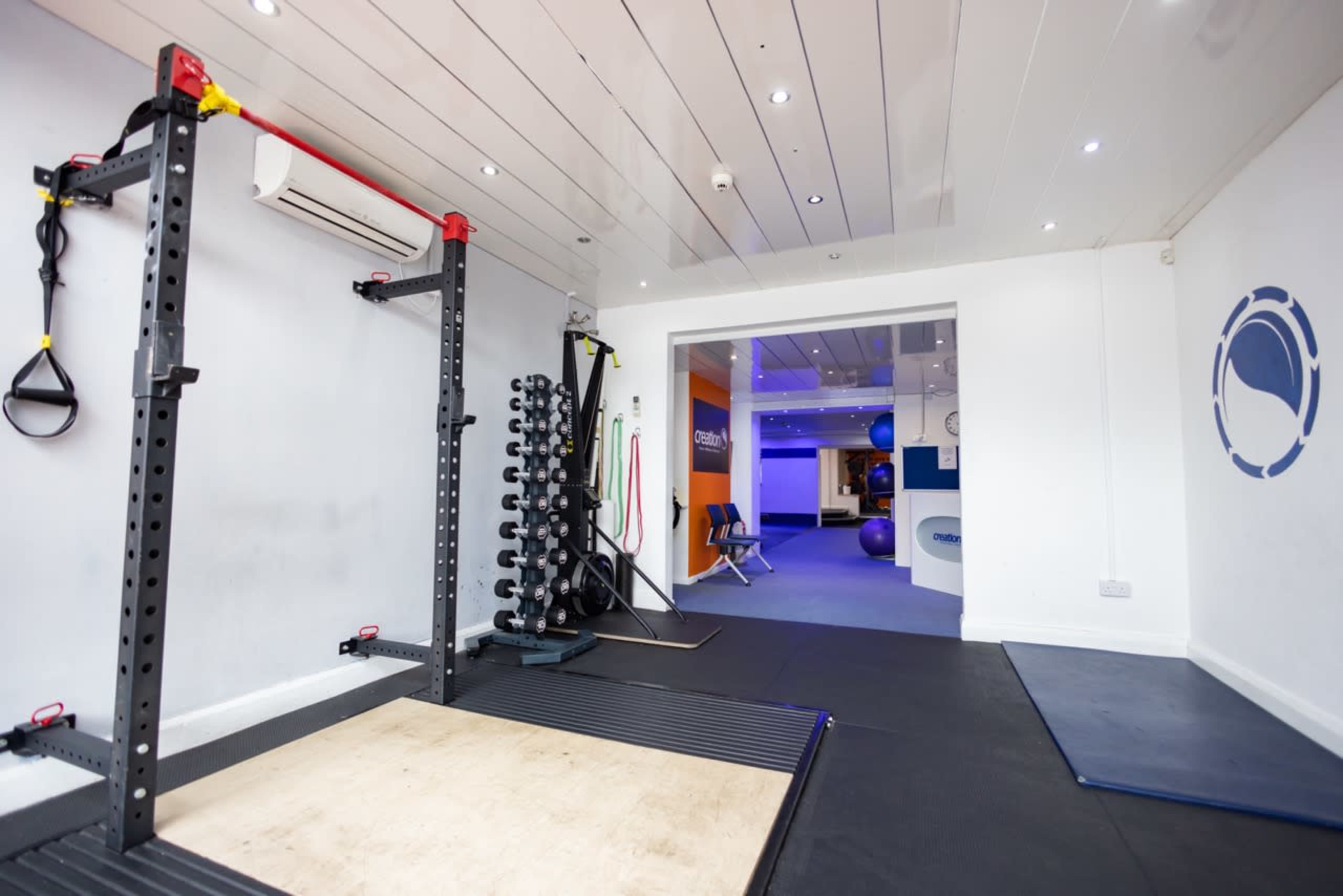 The image shows a sparse gym interior featuring a power rack, a set of dumbbells, and a doorway leading to another room.