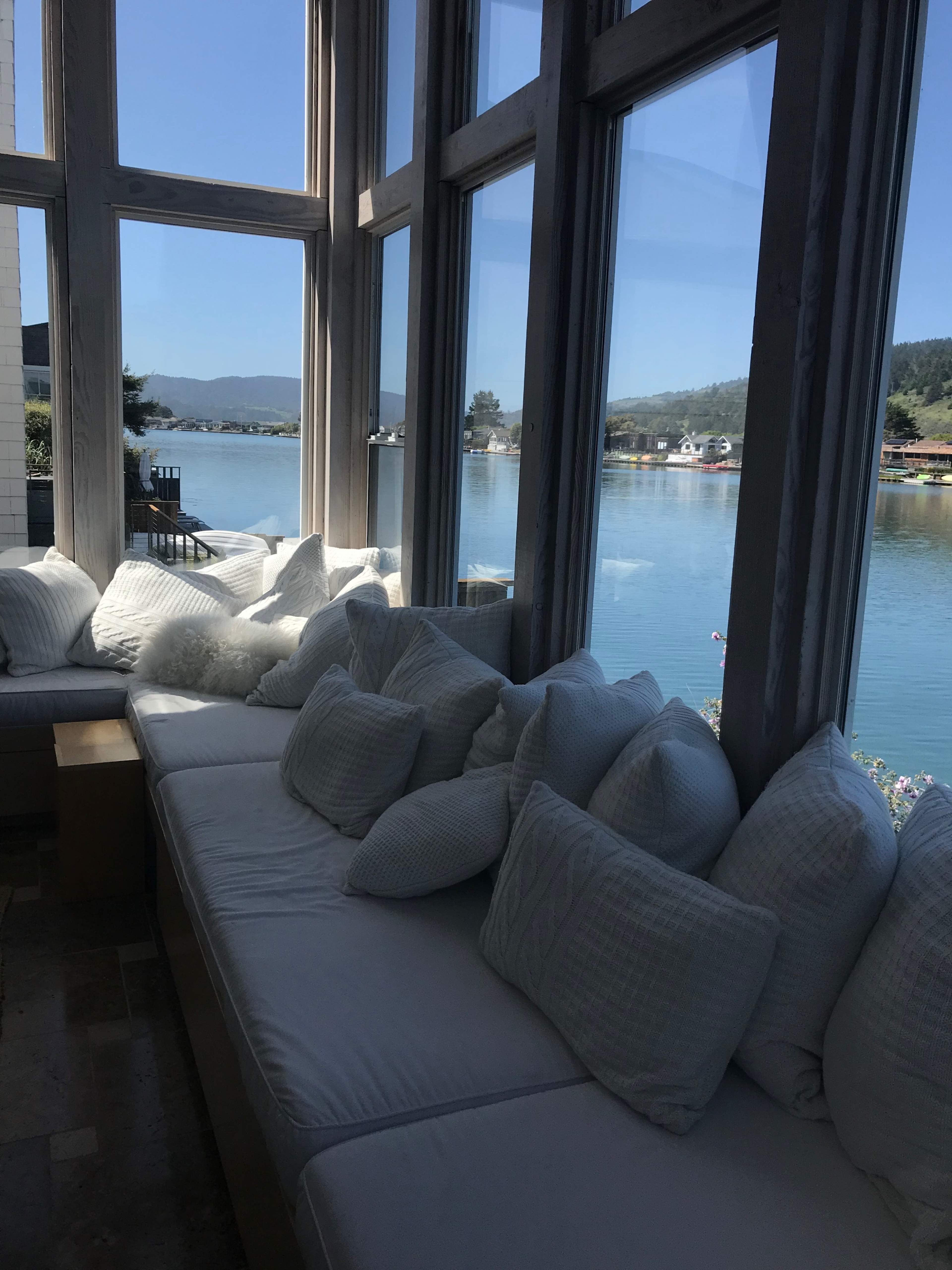 A cozy seating area with white cushions overlooks a calm lake and distant hills through large windows.