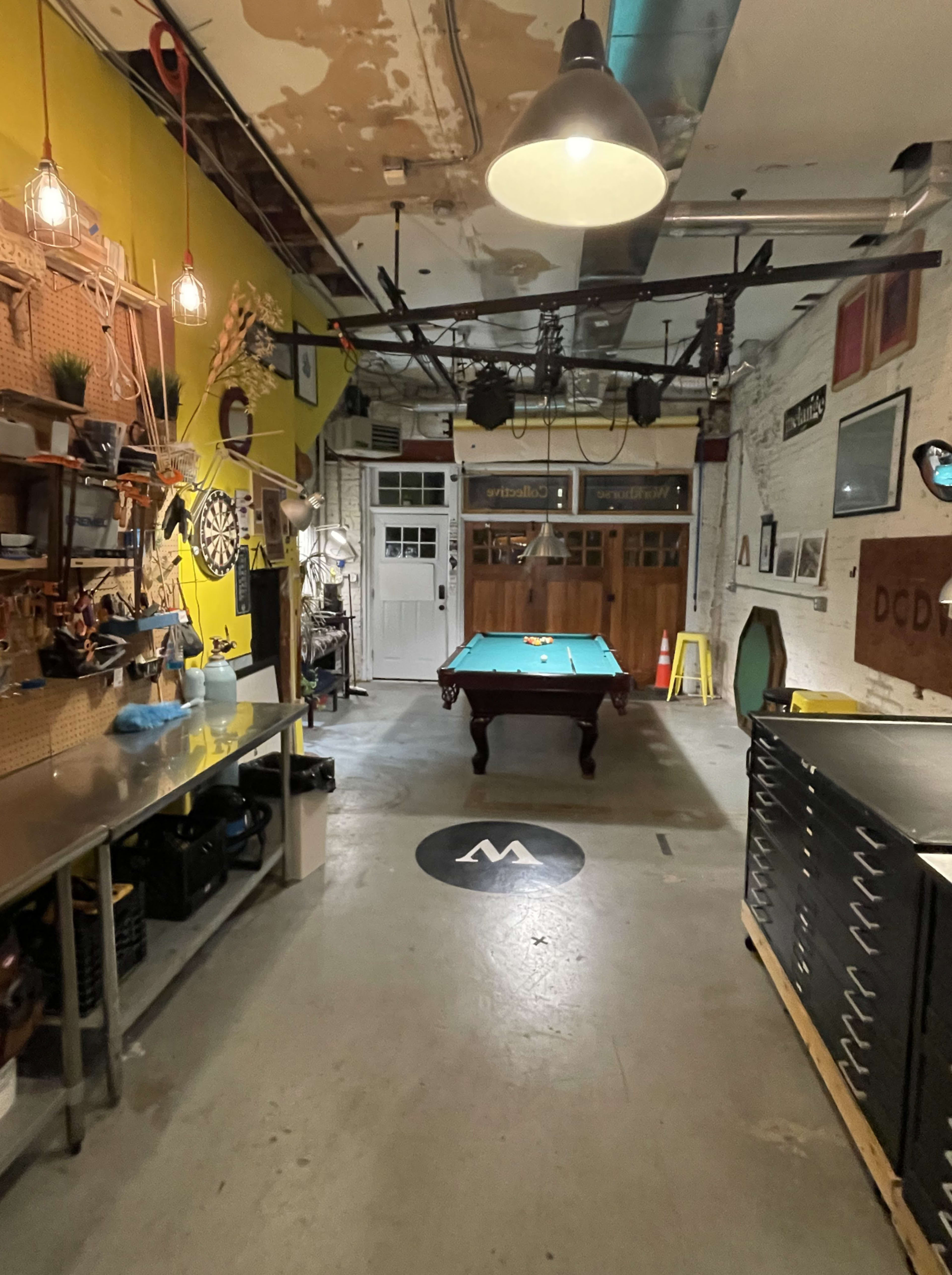 The image shows a spacious workshop or game room with a green pool table centered on a concrete floor, surrounded by various tools and equipment against the walls.