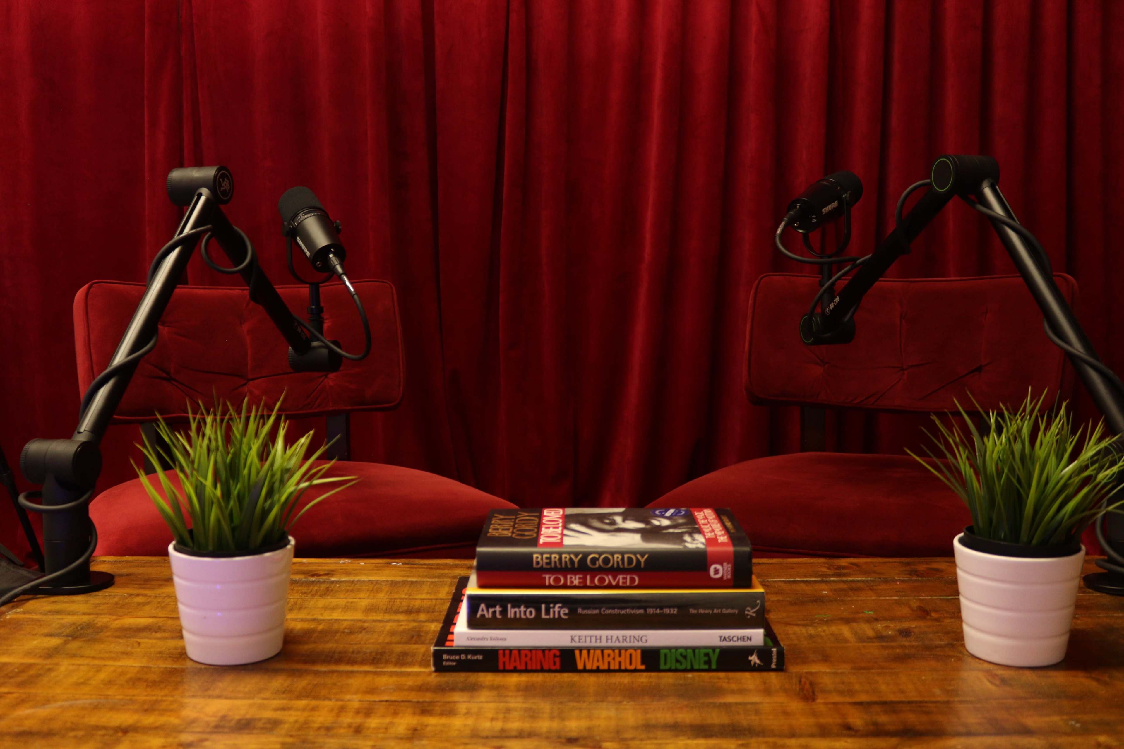 A wooden table with stacked books and small potted plants, flanked by two empty red chairs and microphone stands against a red curtain backdrop.