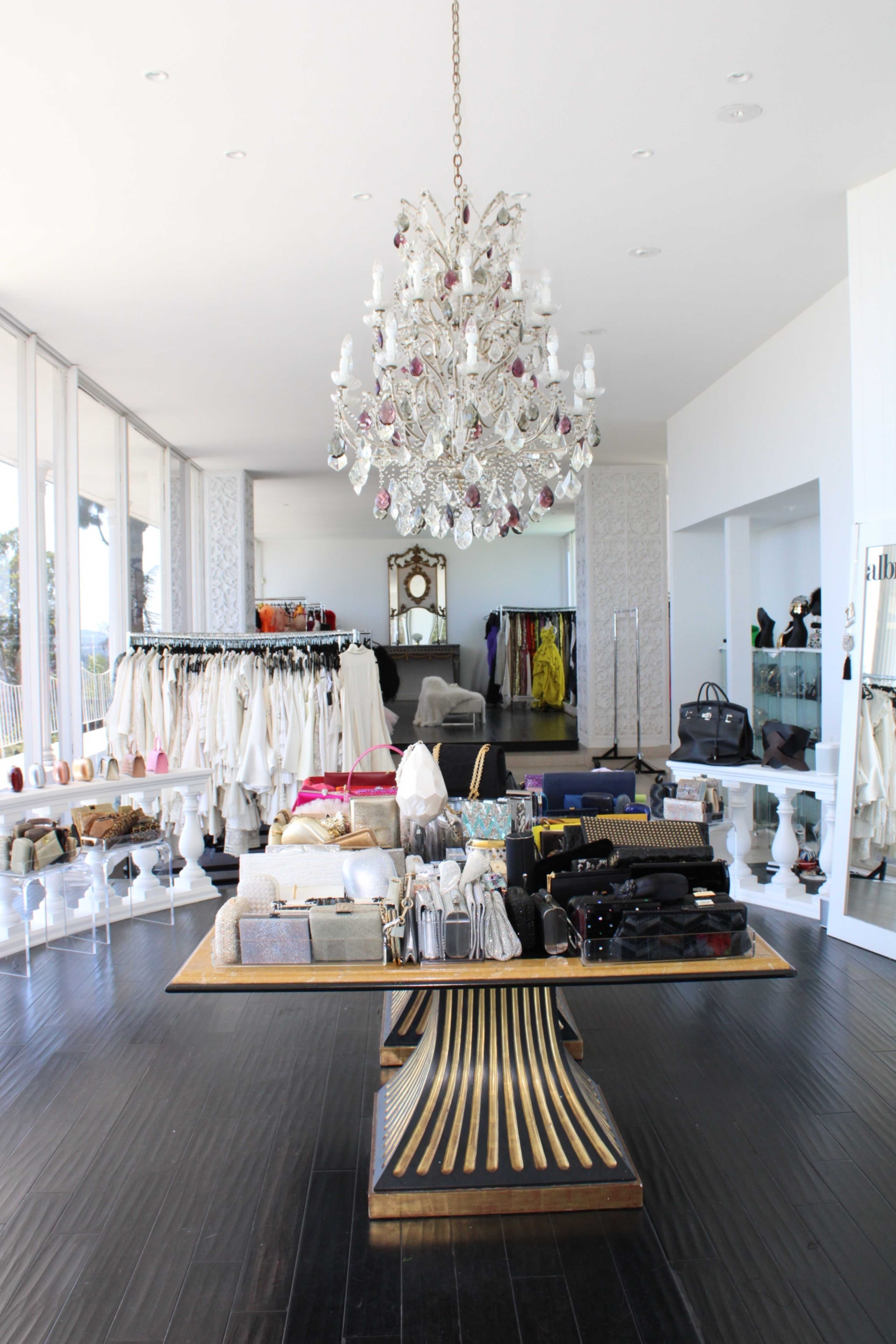The image shows a modern retail clothing store featuring a central display table, a chandelier overhead, and racks filled with various garments in the background.