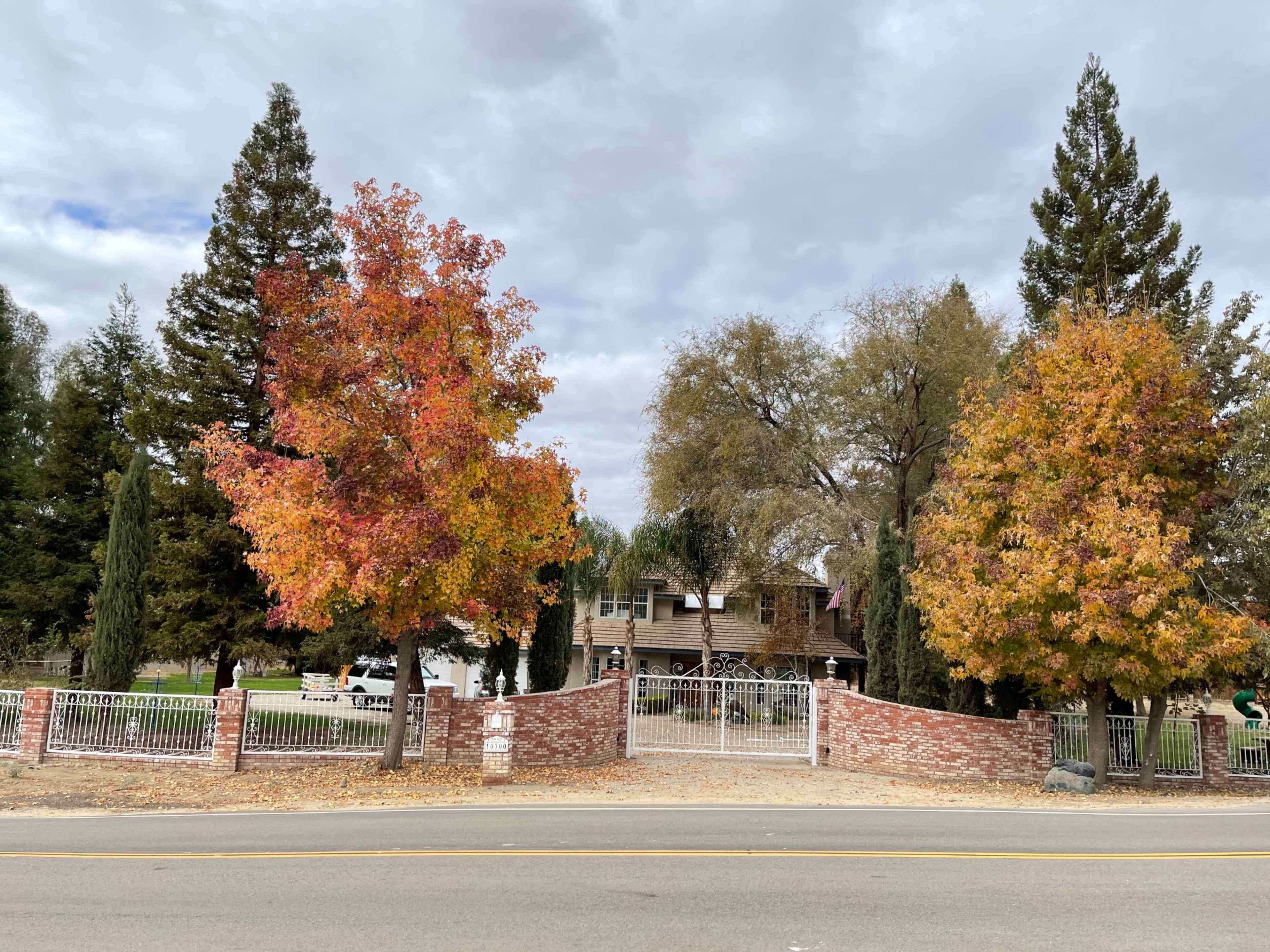 A two-story house with a wooden facade is set behind a brick fence and surrounded by trees displaying autumn foliage.