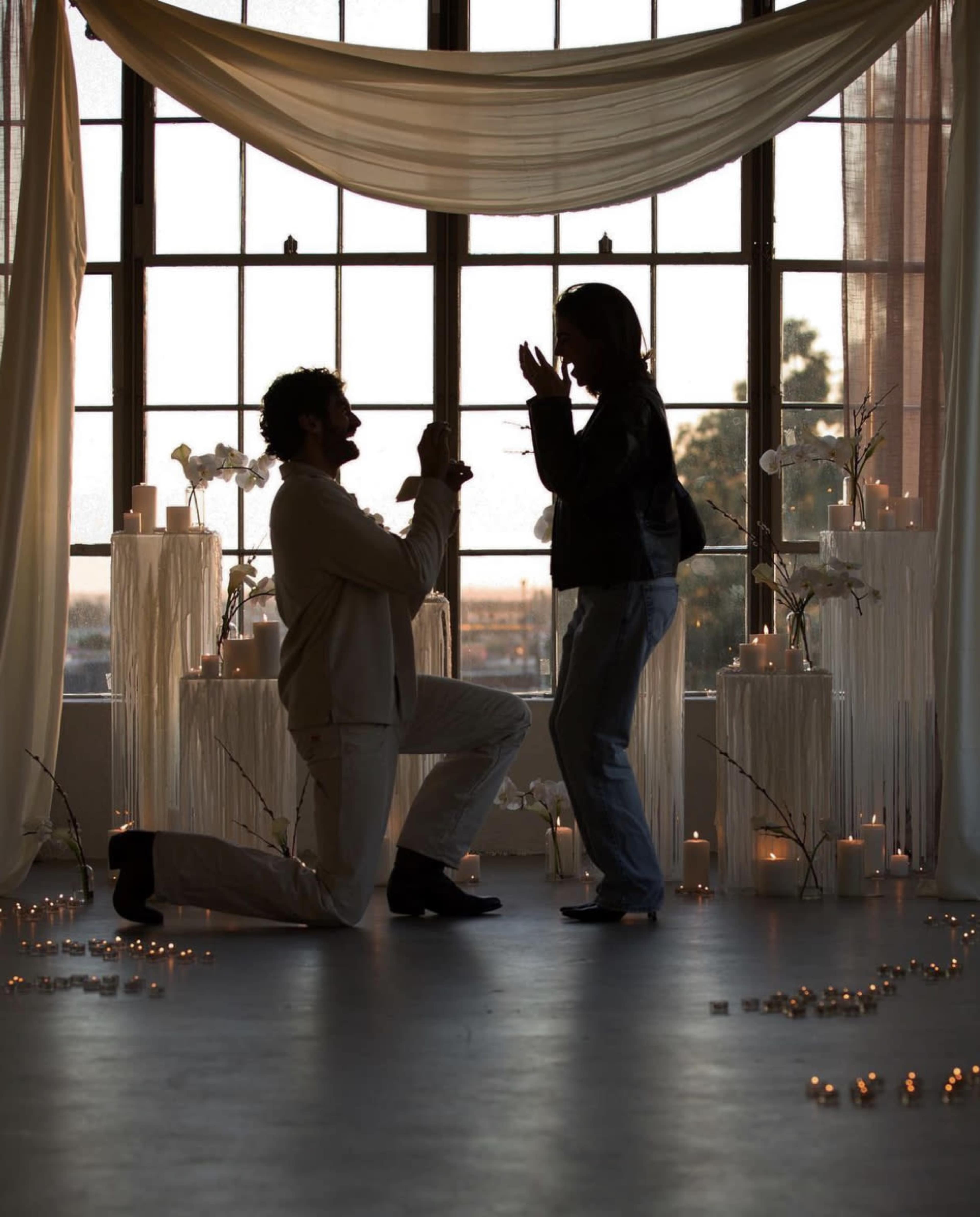 A man kneels with a ring in hand while proposing to a woman in a softly lit room decorated with candles and flowers.