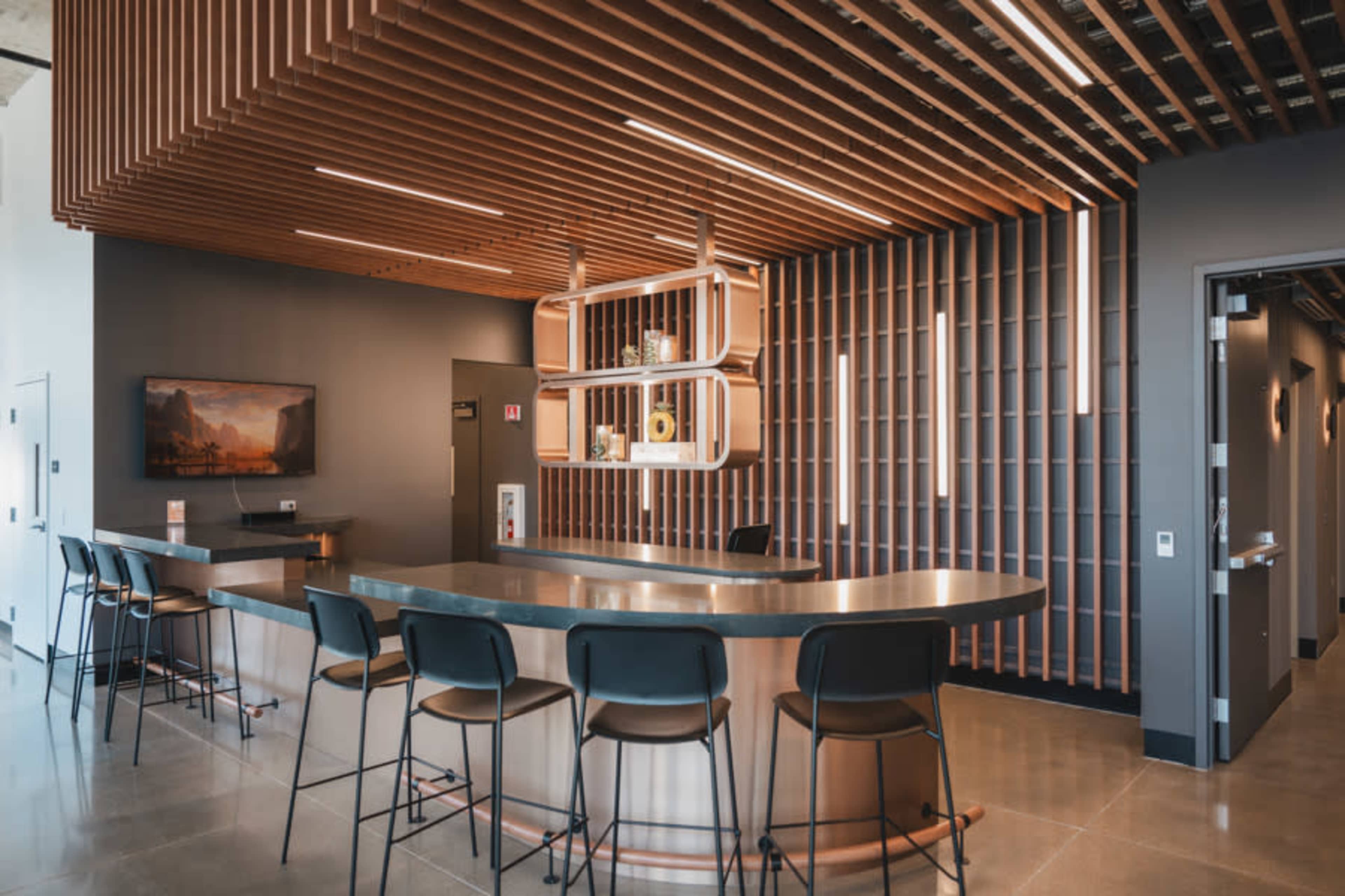 The image shows a modern reception area with a curved metal counter, wooden slat ceiling, and sleek black barstools.