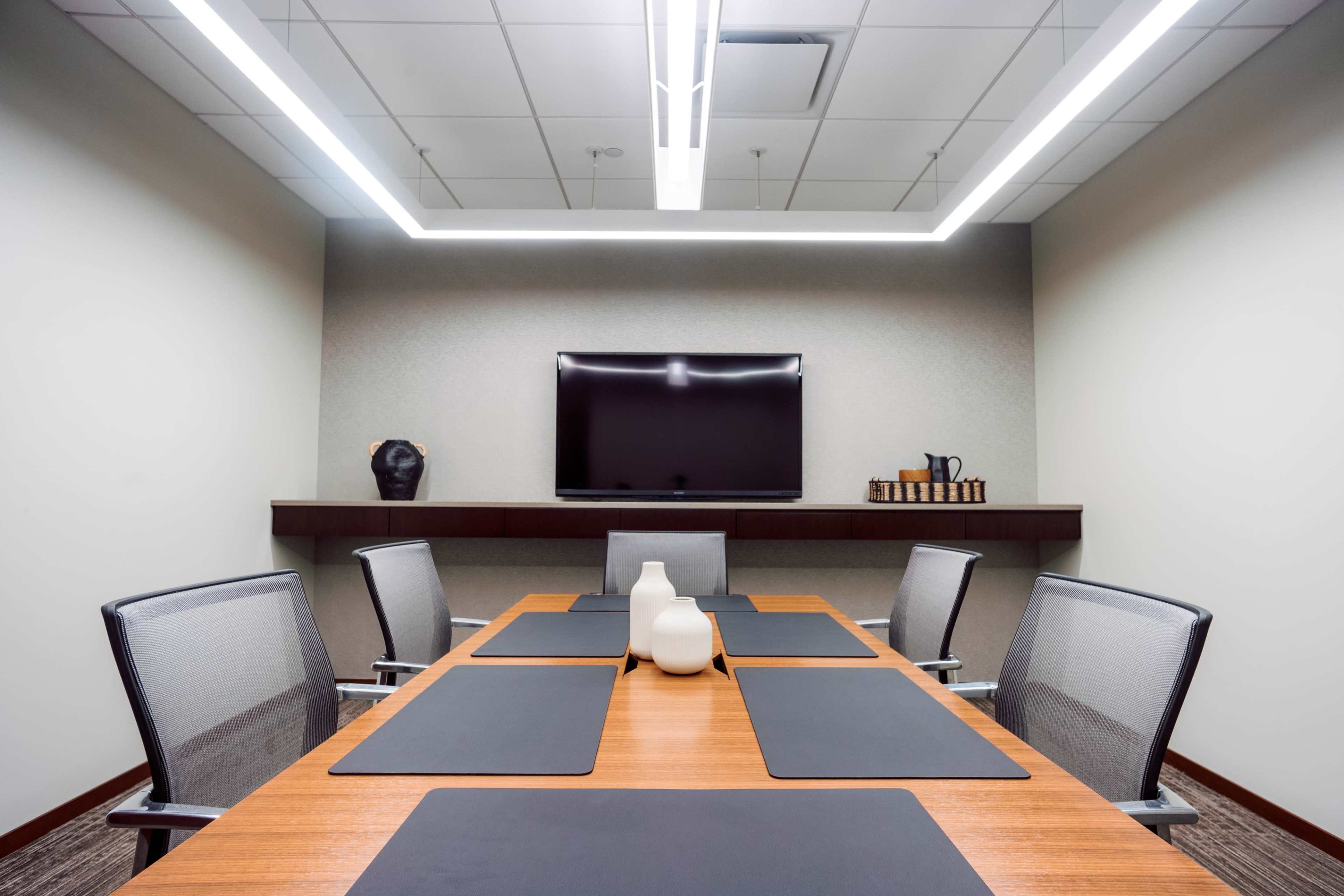 A modern conference room features a wooden table surrounded by mesh chairs, with a wall-mounted television and decorative items on a shelf.