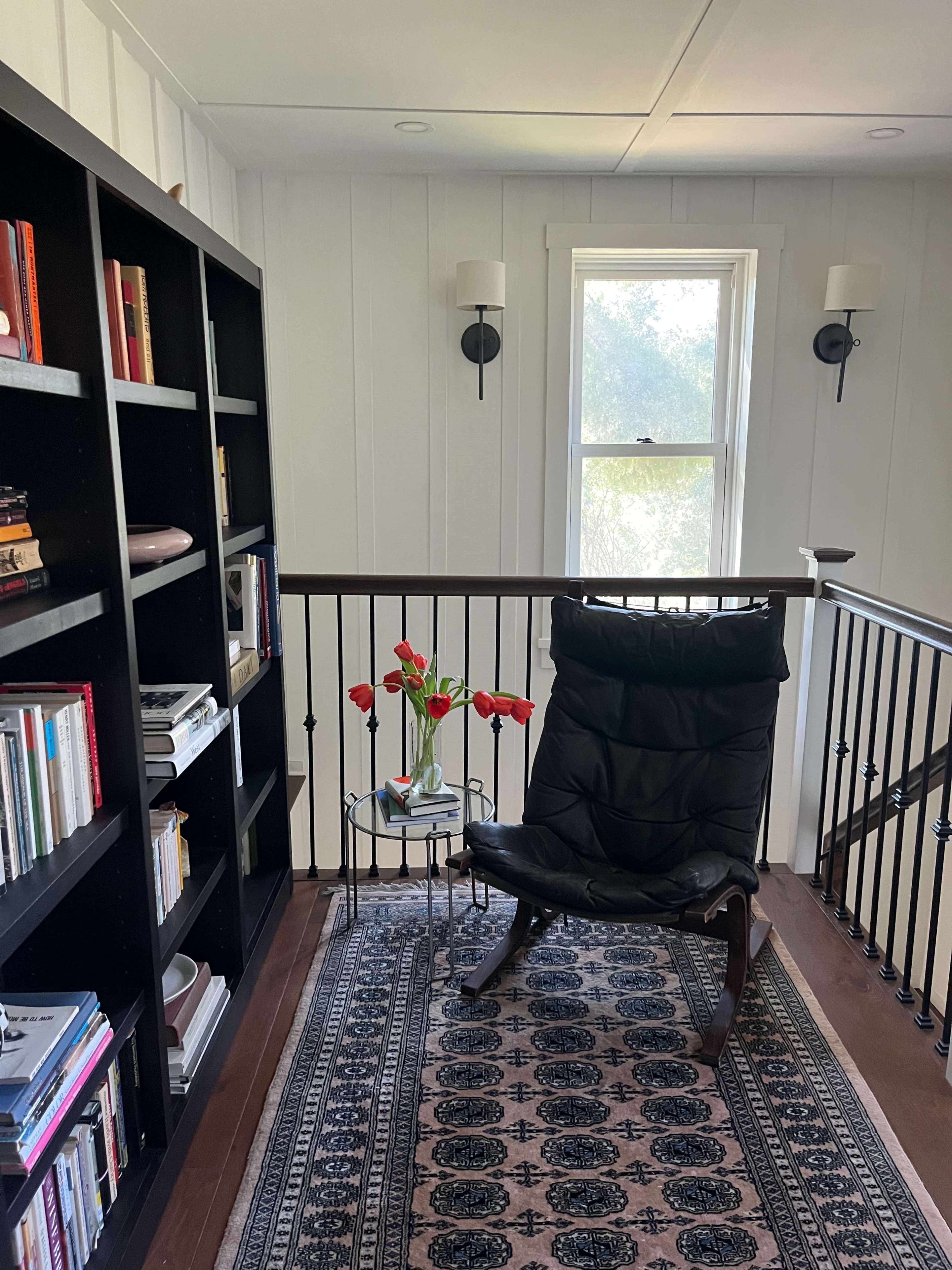 A black chair sits beside a small table with a vase of red flowers, adjacent to a bookshelf and a window in a well-lit room.