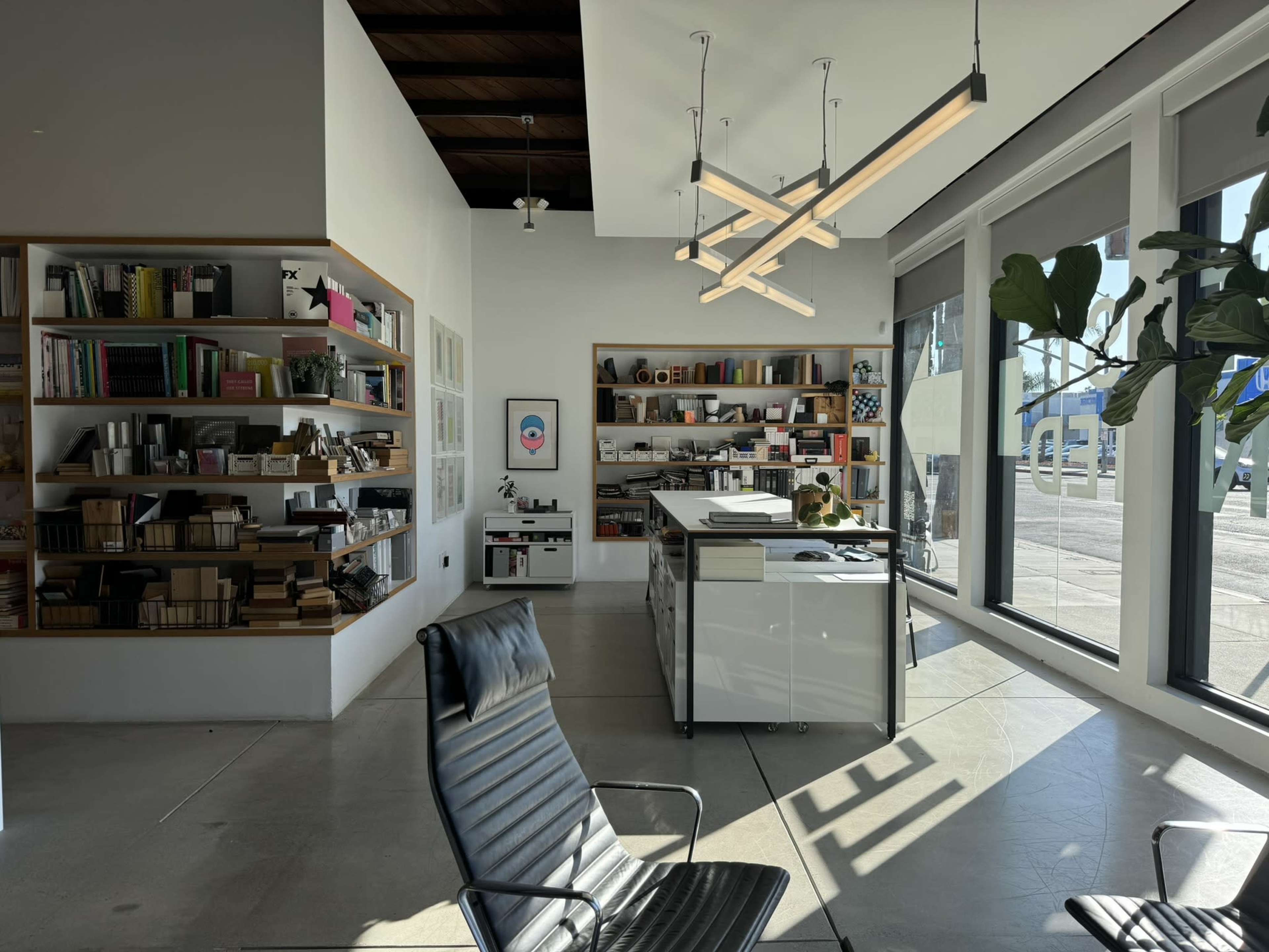 The image shows a modern office interior with a sleek chair, a bookshelf filled with various items, and large windows allowing natural light to enter the space.