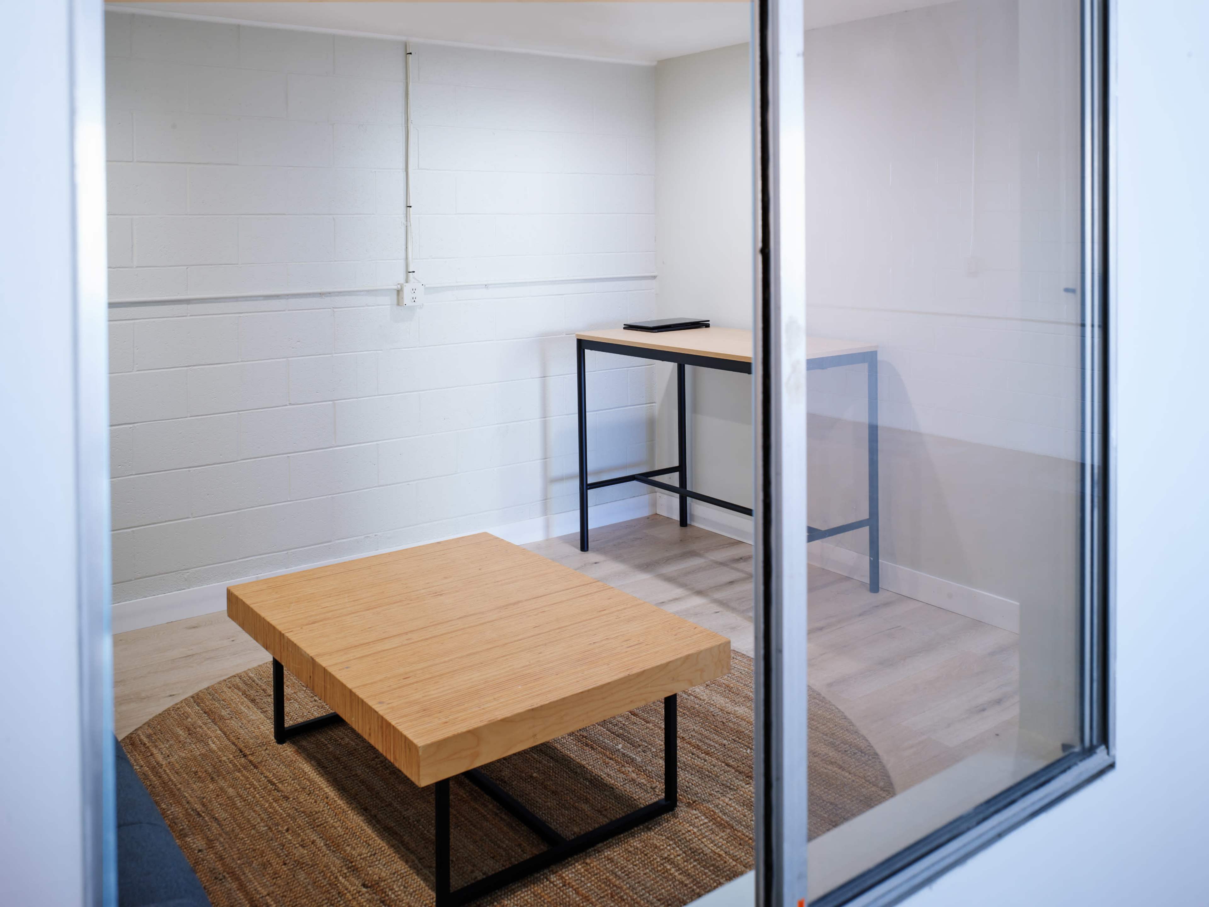 A minimalistic room with a wooden table, a round rug, and a desk viewed through a glass wall.