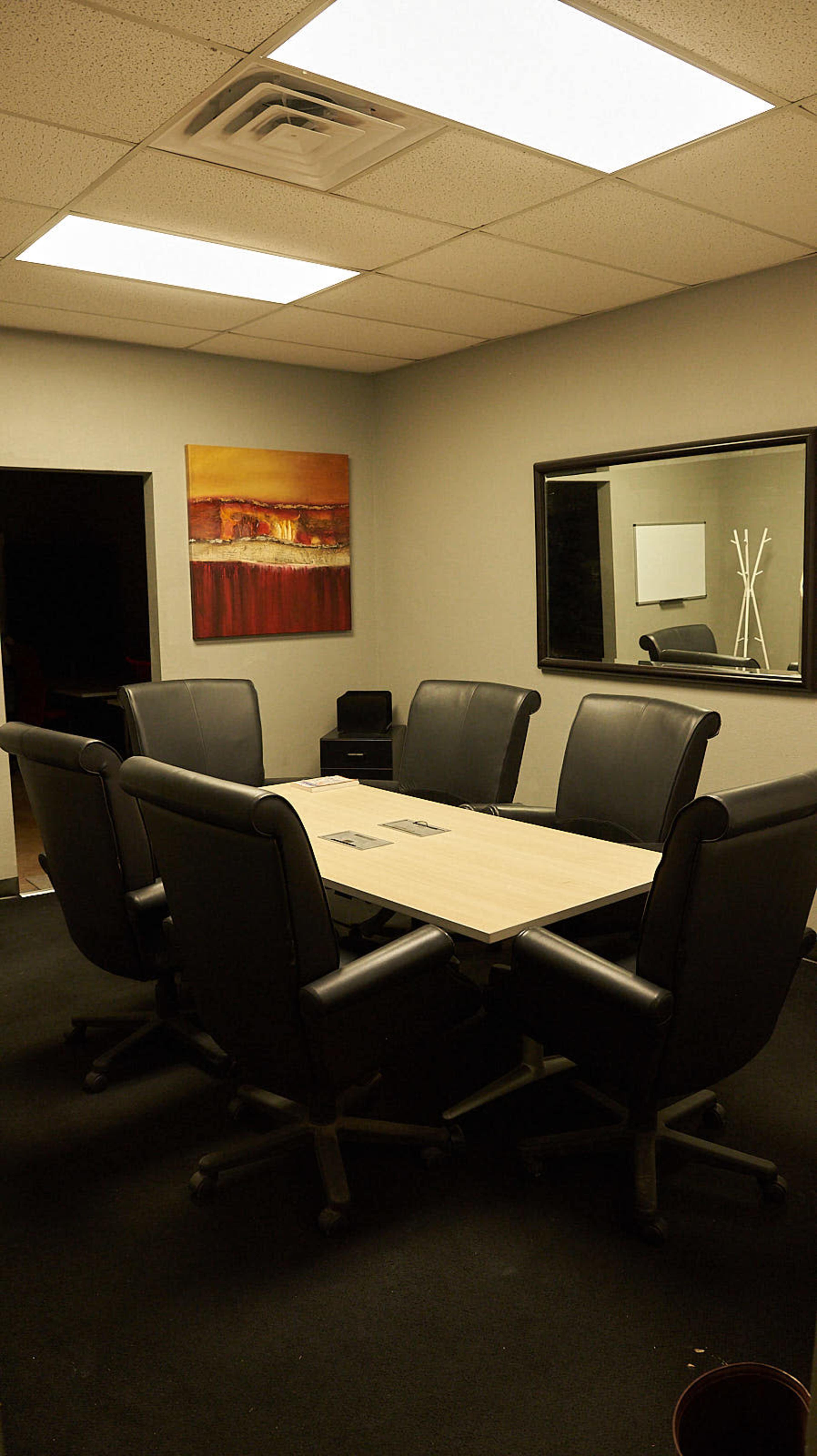 A conference room features a large table surrounded by six black leather chairs, with two artworks on the walls and a mirror opposite the entrance.