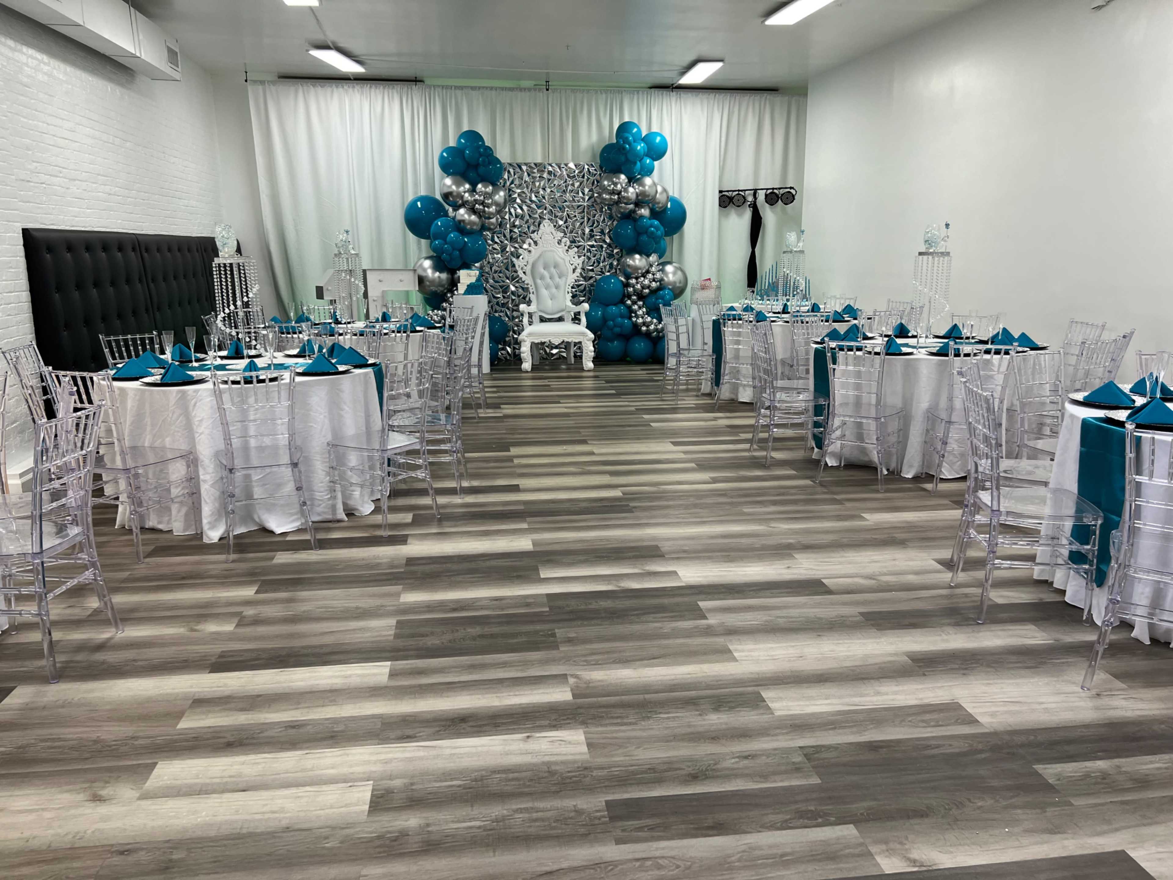 The image shows a decorated event space with round tables covered in white tablecloths and turquoise accents, featuring a backdrop of blue and silver balloons.