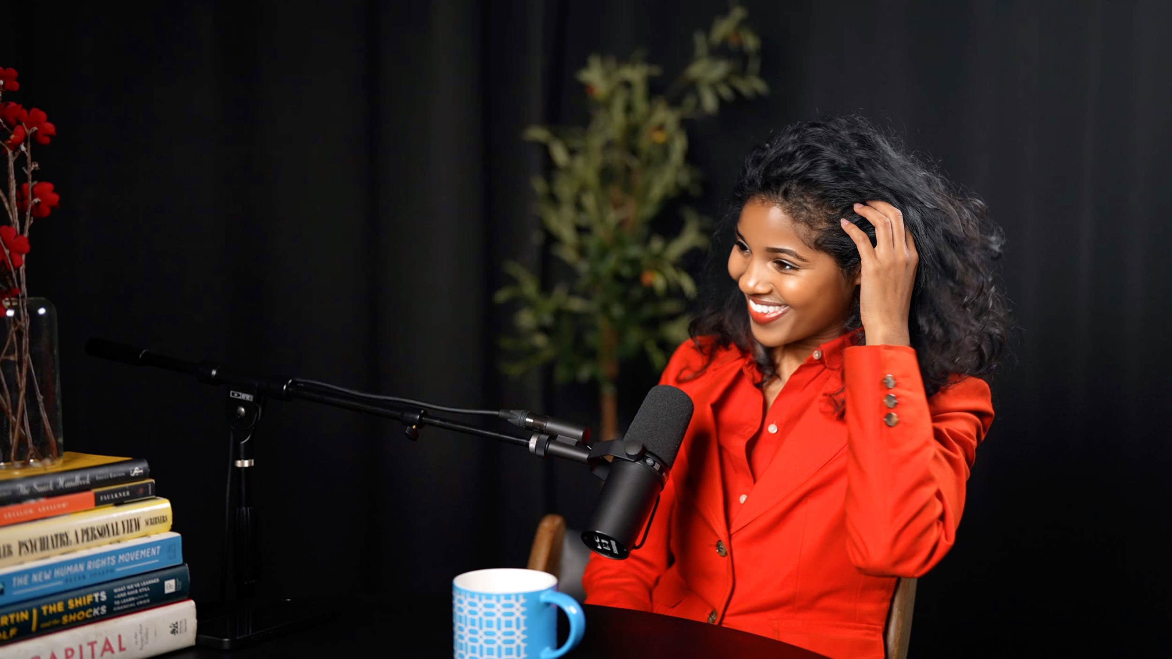 A woman wearing a red blazer smiles while seated at a table with a microphone and books stacked beside her.
