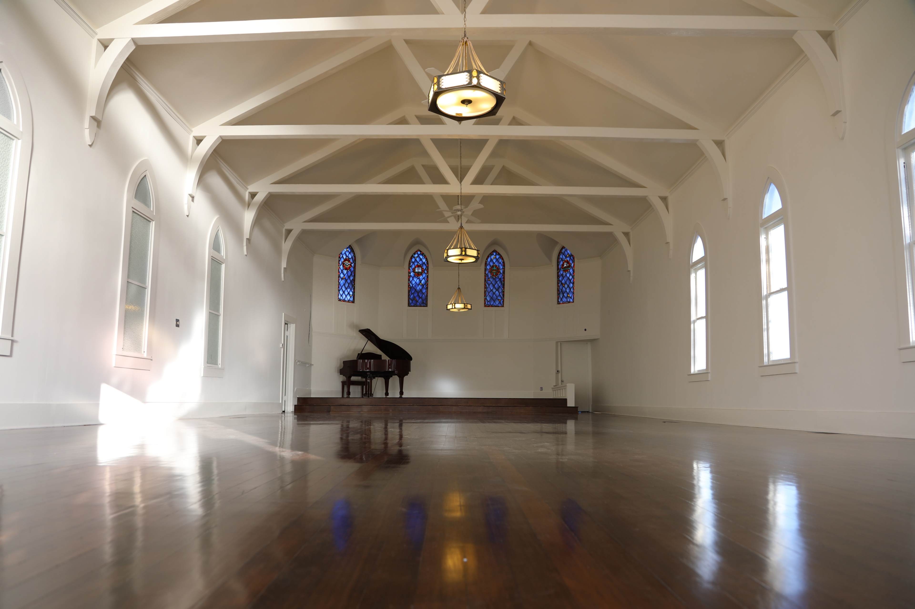 A spacious, well-lit room with wooden floors, large windows, and a grand piano on a raised platform.